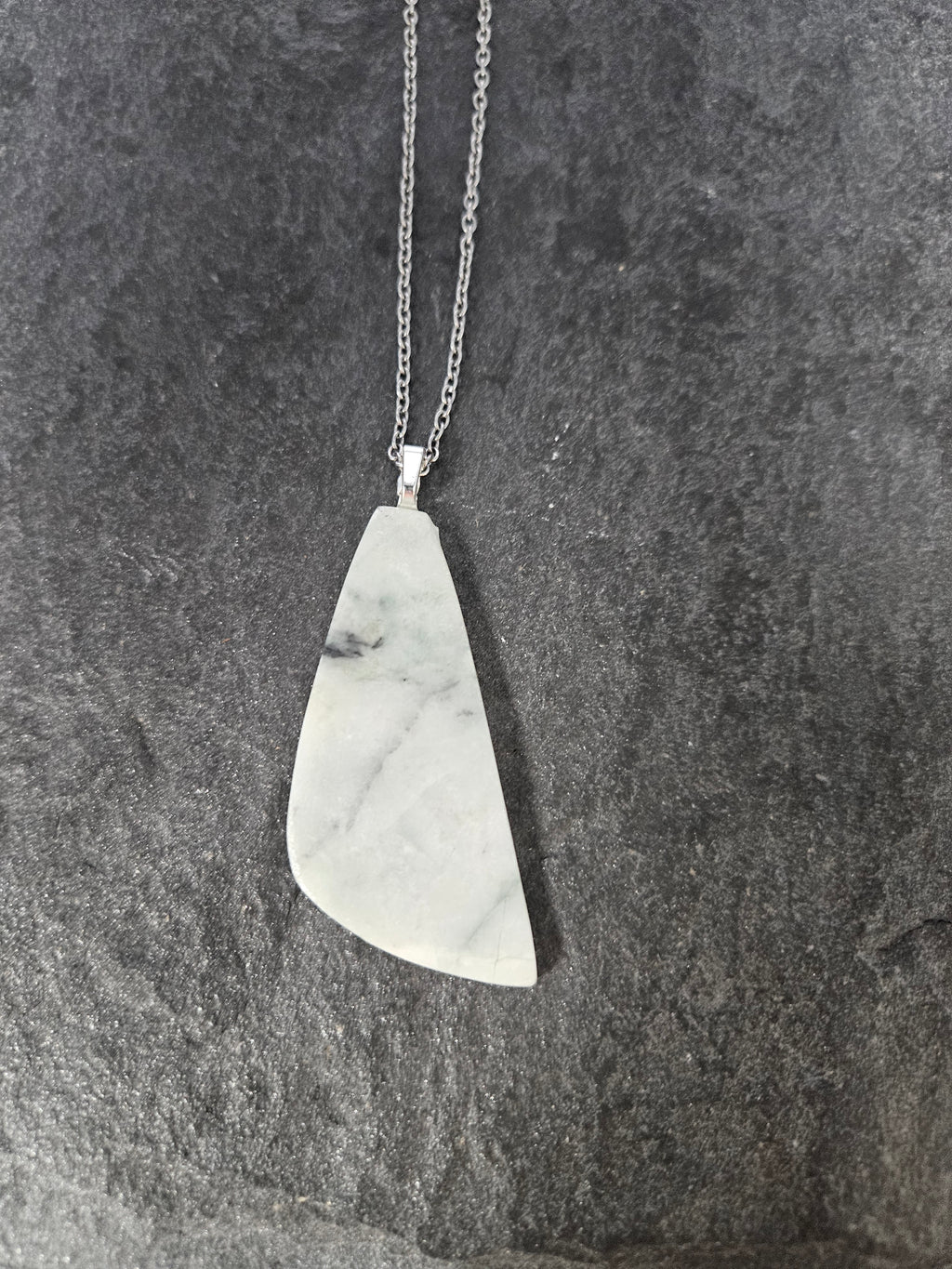 A silver pendant necklace featuring a polished, irregularly shaped white stone with gray veining, displayed on a dark textured slate surface.