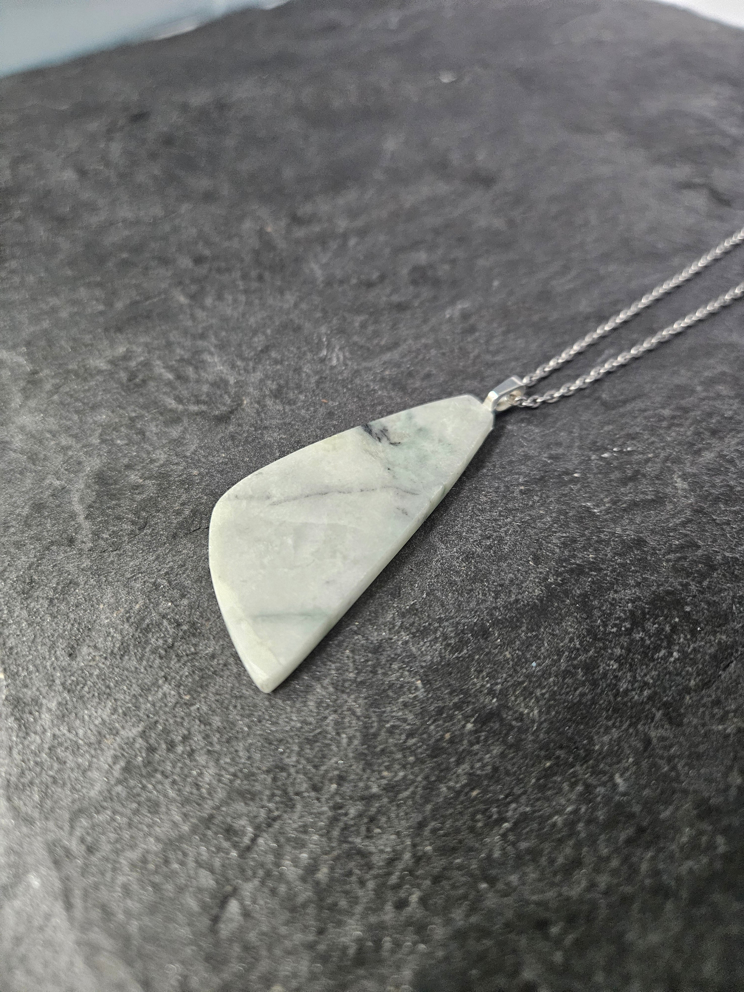 A silver necklace with a polished, irregular triangular pendant made of light gray and white marbled stone, resting on a dark textured surface.