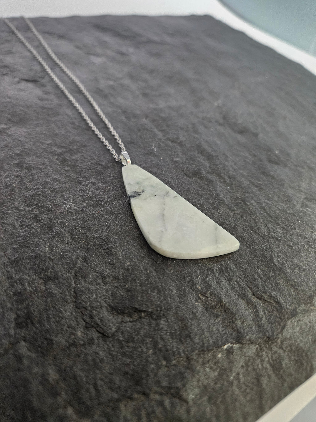 A silver necklace with a triangular pendant made of light gray and white marbled stone, displayed on a dark textured slate surface.