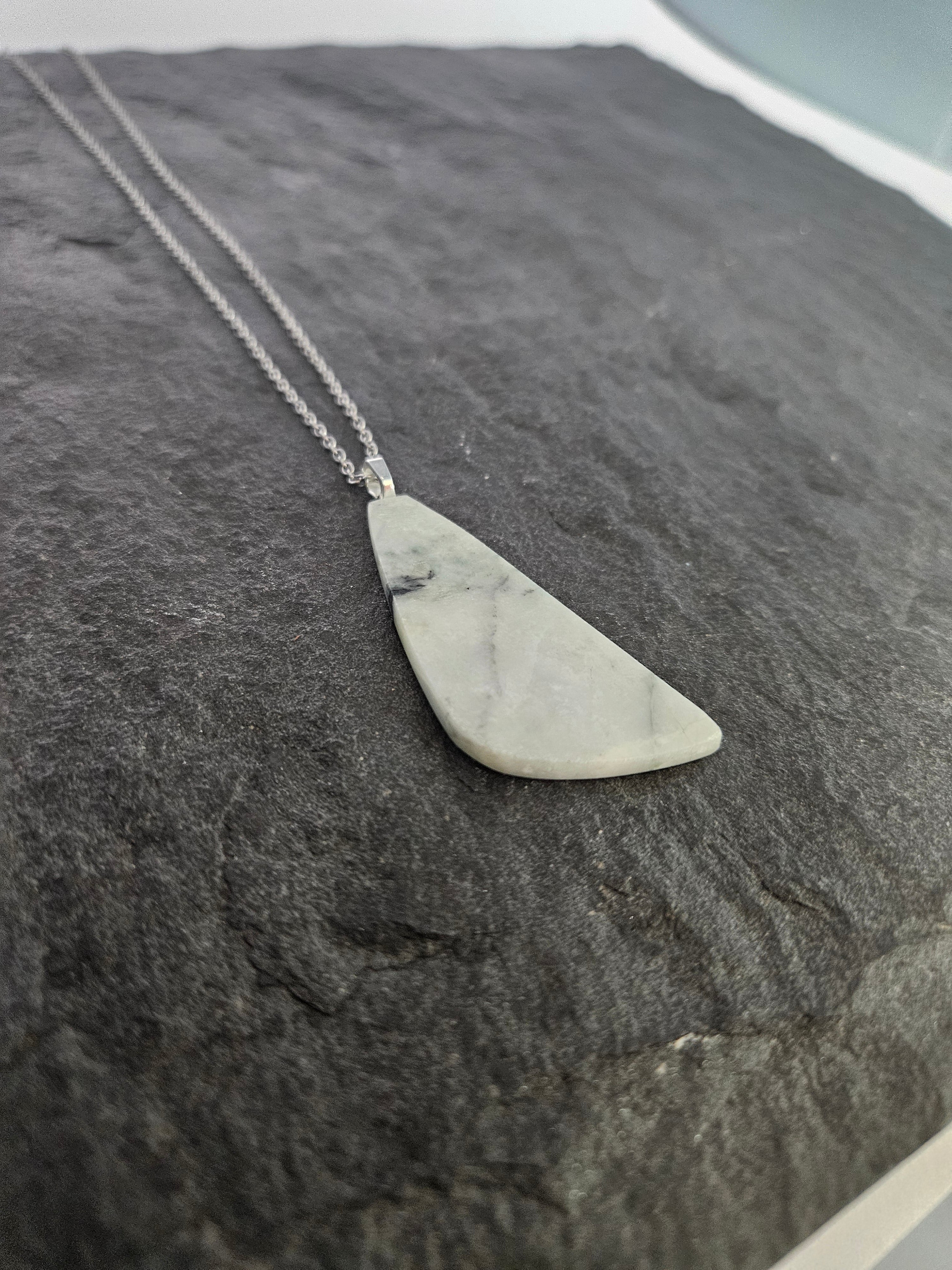 A silver necklace with a triangular pendant made of light gray and white marbled stone, displayed on a dark textured slate surface.