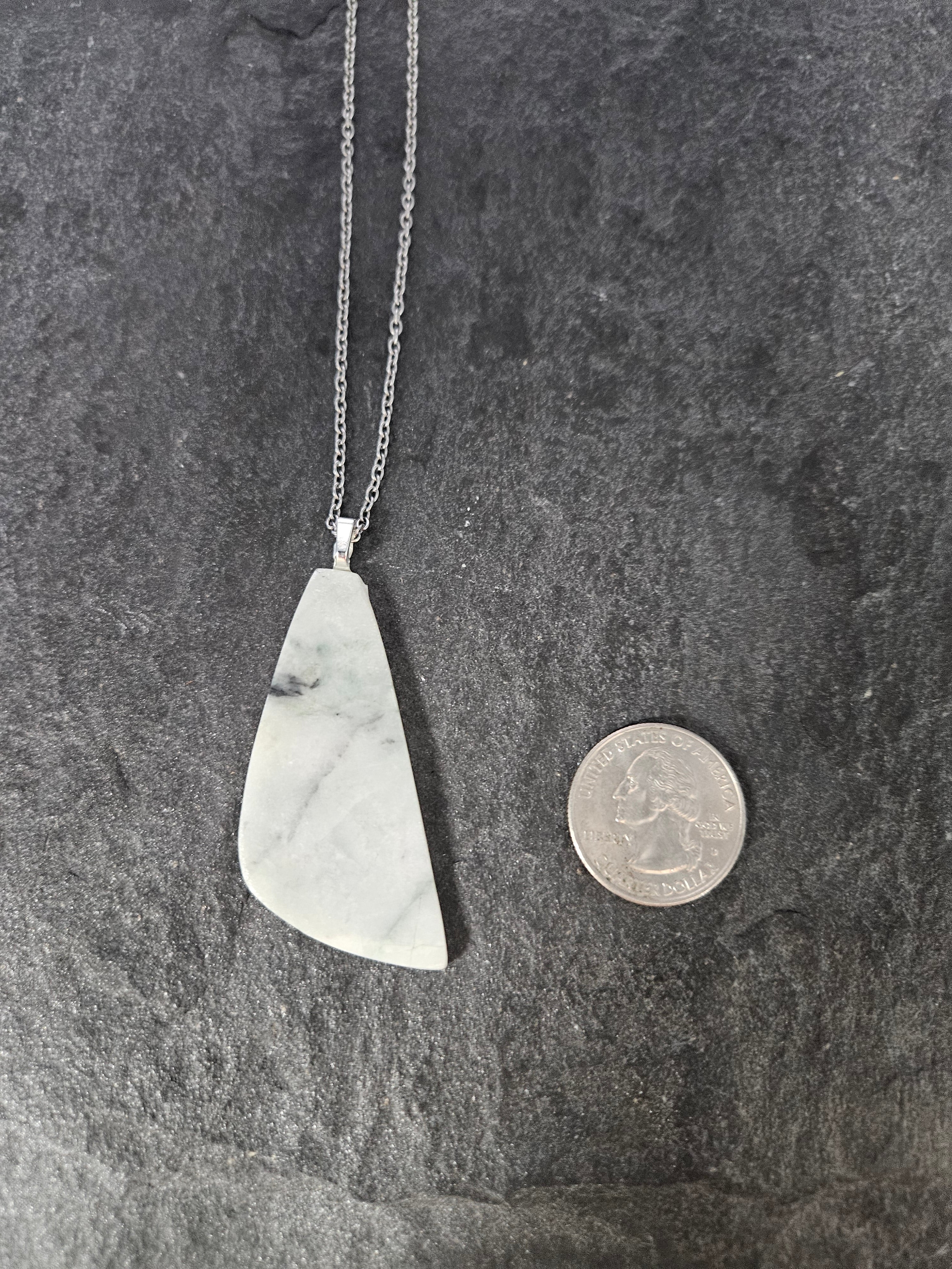 A silver necklace with a teardrop-shaped white and gray marble pendant, displayed next to a US quarter coin to show scale on a dark textured slate background.