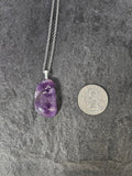 A silver chain necklace with a polished purple teardrop-shaped gemstone pendant, shown next to a US quarter coin for scale on a dark gray textured surface.
