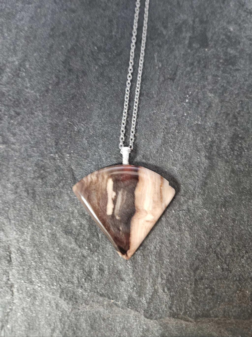 A silver chain necklace with a triangular pendant made from a polished wood slice showing natural brown and beige grain, displayed on a dark gray slate surface.