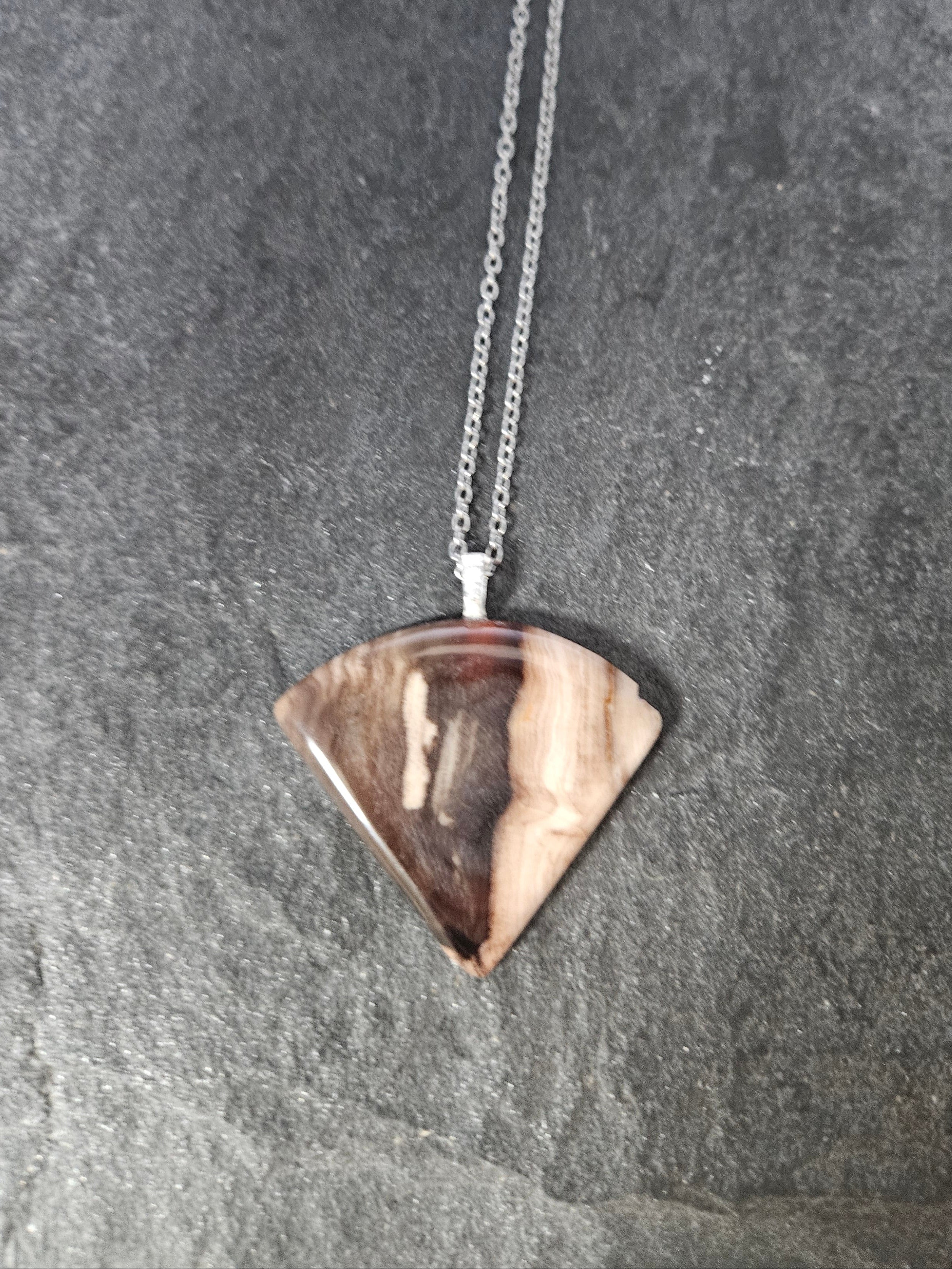 A silver chain necklace with a triangular pendant made from a polished wood slice showing natural brown and beige grain, displayed on a dark gray slate surface.