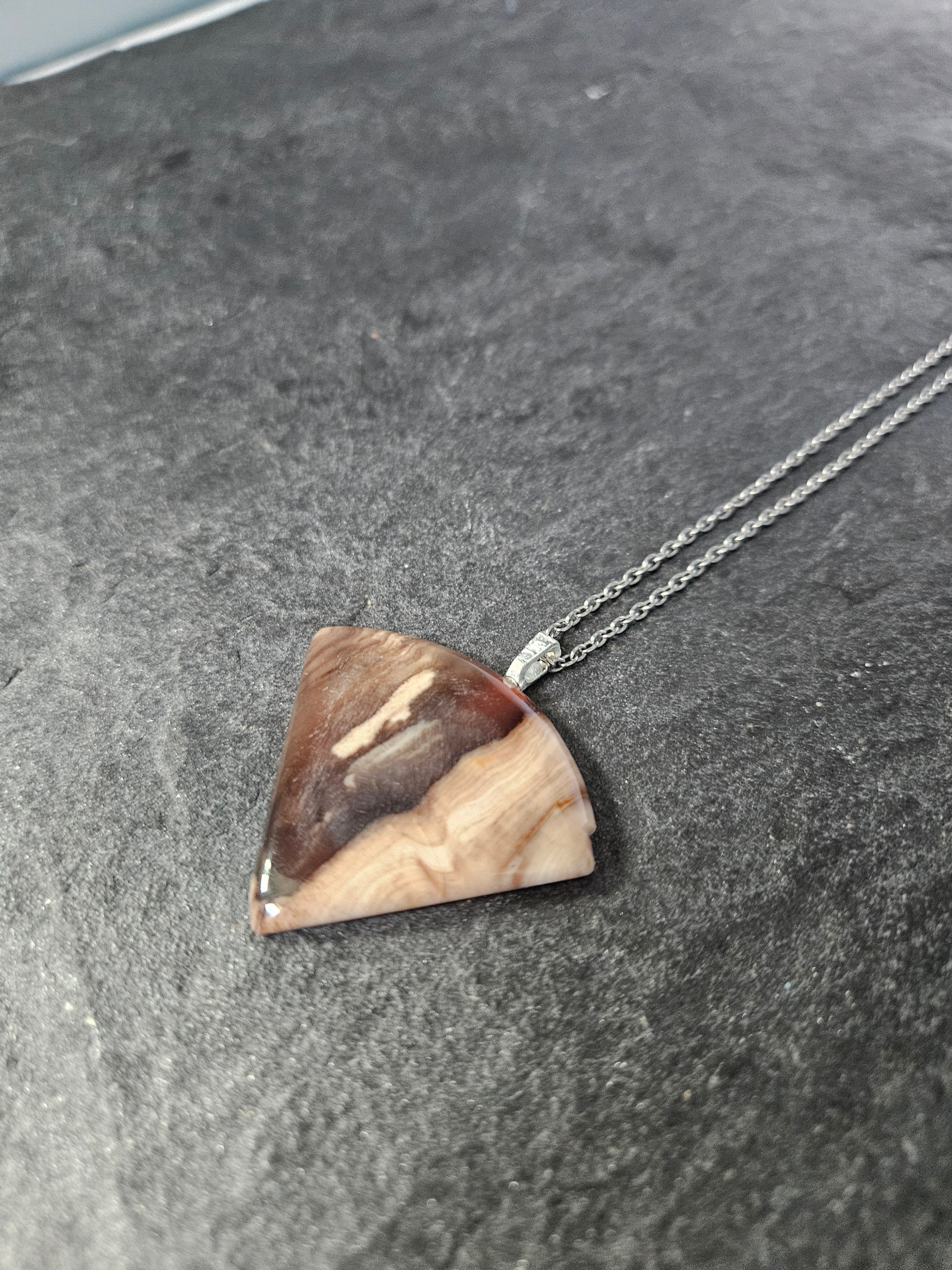 A triangular pendant necklace made of polished brown and cream stone with a silver chain, displayed on a dark textured slate surface.