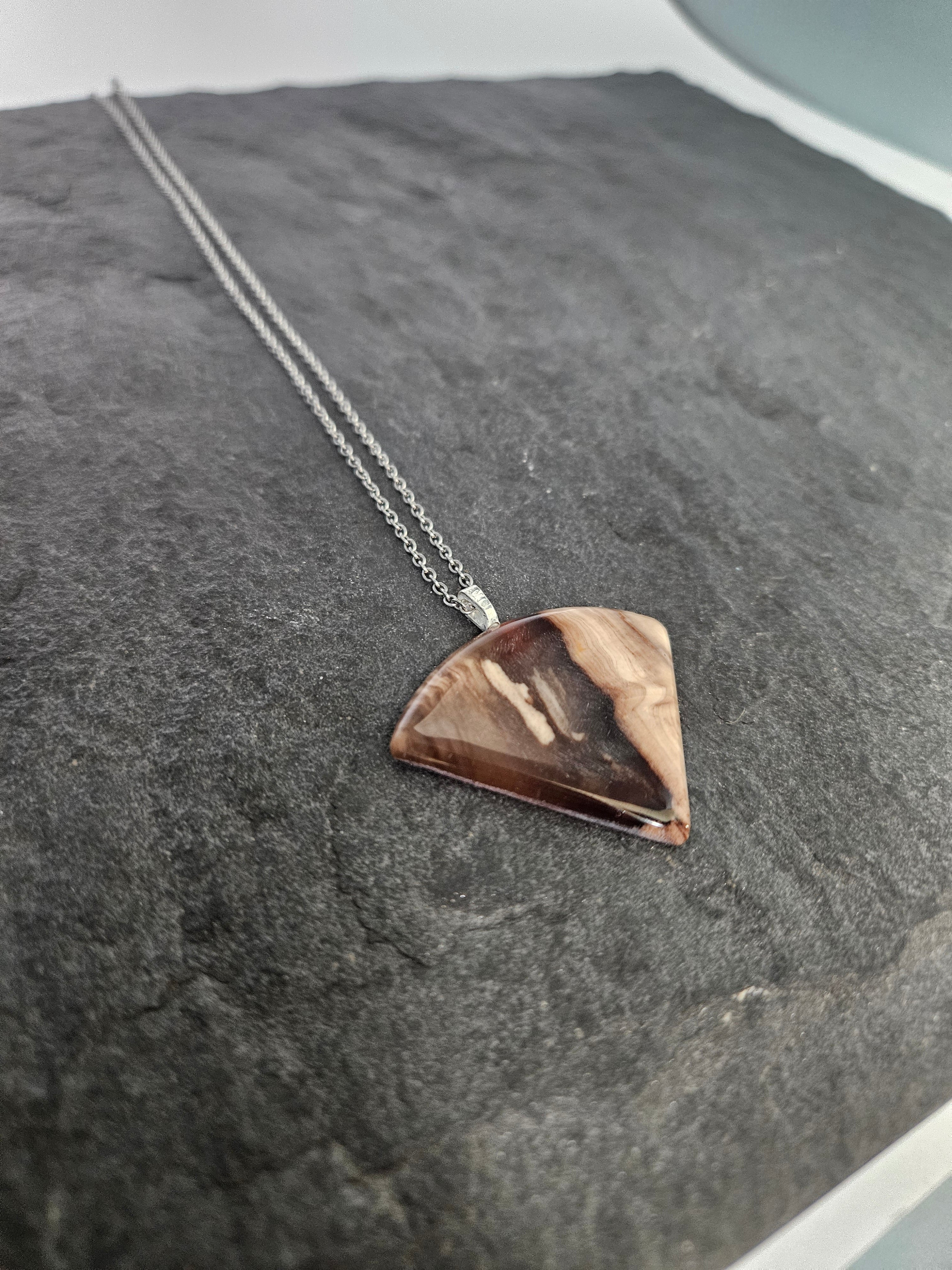A silver chain necklace with a wedge-shaped pendant made from polished brown wood with natural grain patterns, displayed on a dark textured slate surface.