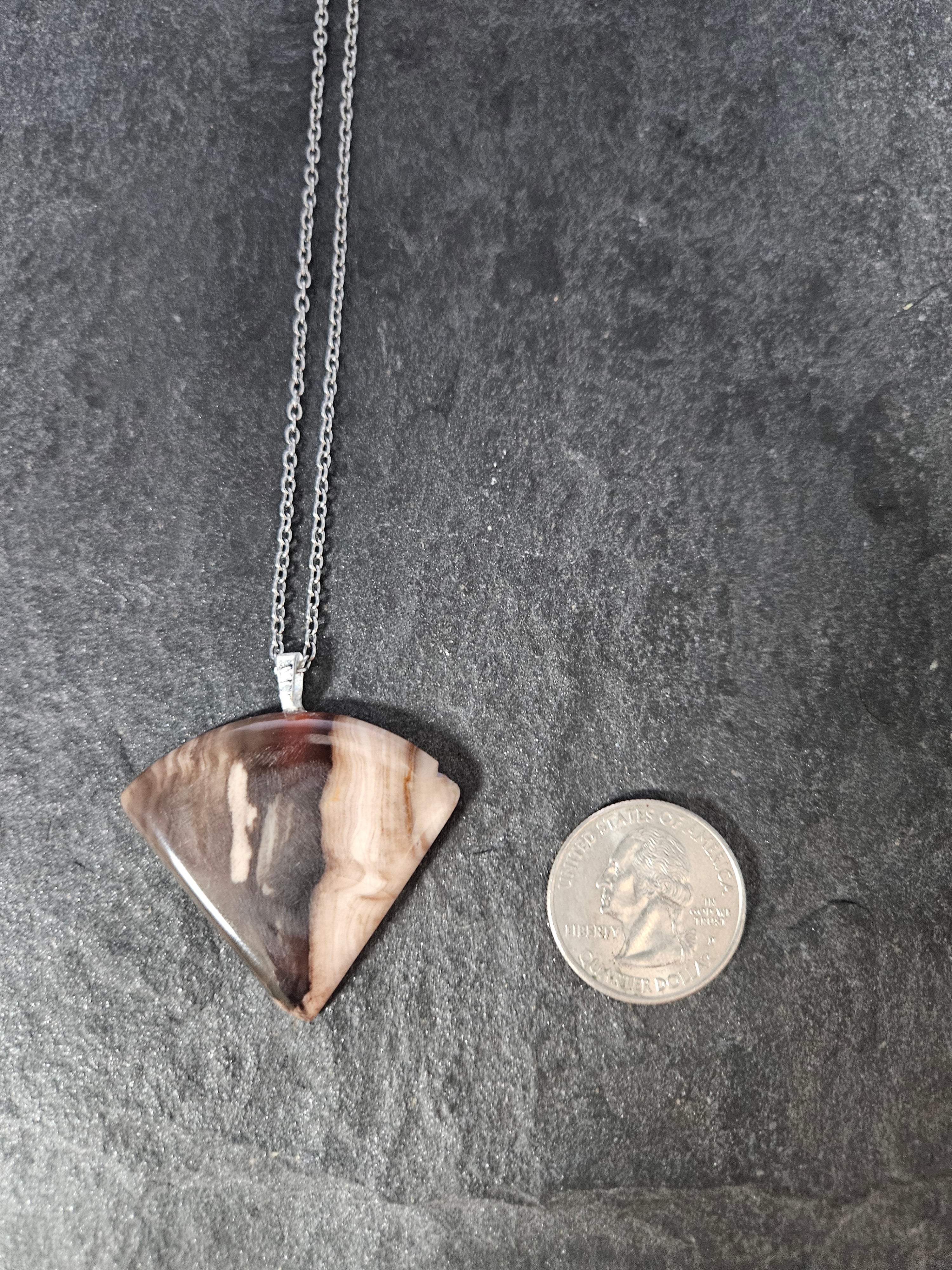 A silver chain necklace with a triangular pendant made of natural brown and beige agate, displayed next to a US quarter coin to show its size on a dark gray slate background.