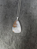 A silver chain necklace with a polished, irregular-shaped pendant made of white and gray agate featuring natural brown and orange mineral inclusions, displayed on a dark textured slate surface.