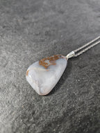 A silver pendant necklace featuring a polished, irregular-shaped gray stone with natural brown inclusions, resting on a textured gray surface.