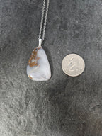 A silver pendant necklace with a raw, polished stone featuring white, gray, and brown natural patterns, displayed next to a US quarter coin for scale on a dark textured background.