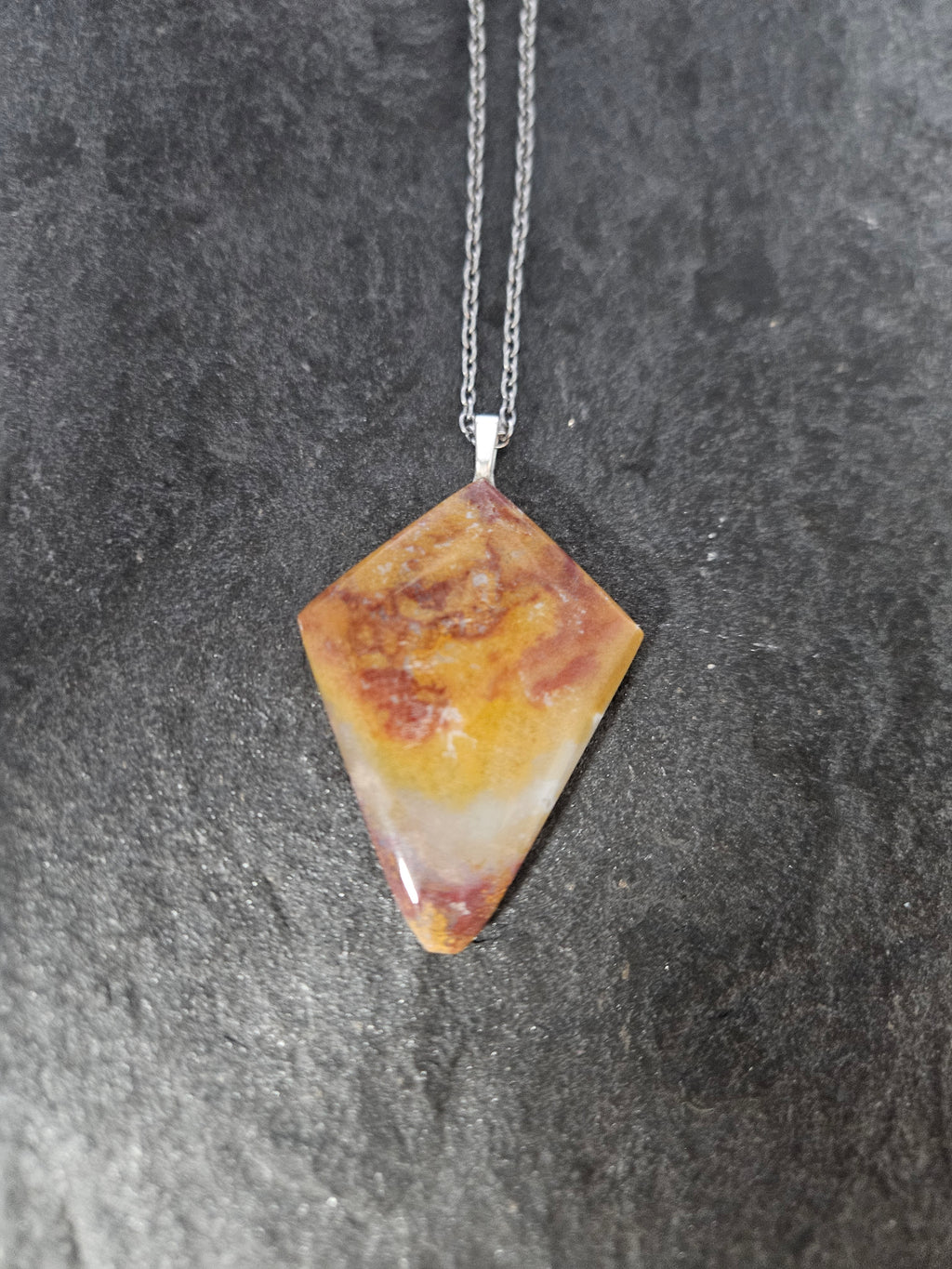 A diamond-shaped gemstone pendant necklace with a marbled pattern of orange, yellow, and reddish-brown, suspended from a silver chain against a dark textured slate background.