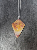 A diamond-shaped gemstone pendant necklace with a marbled pattern of orange, yellow, and reddish-brown, suspended from a silver chain against a dark textured slate background.