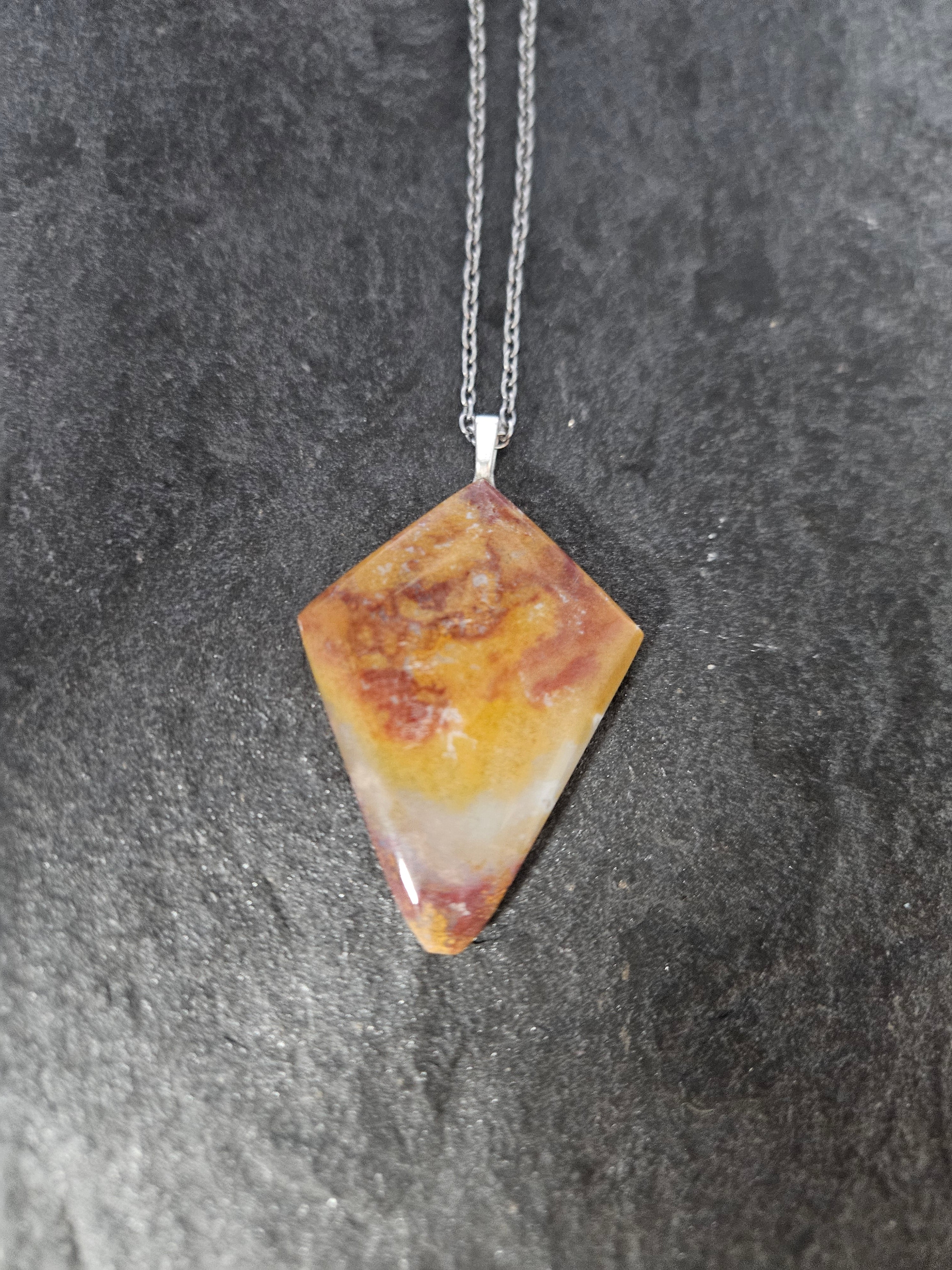 A diamond-shaped gemstone pendant necklace with a marbled pattern of orange, yellow, and reddish-brown, suspended from a silver chain against a dark textured slate background.