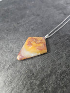 A geometric triangular pendant necklace made of marbled yellow, orange, and white stone with a silver chain, resting on a dark textured slate surface.