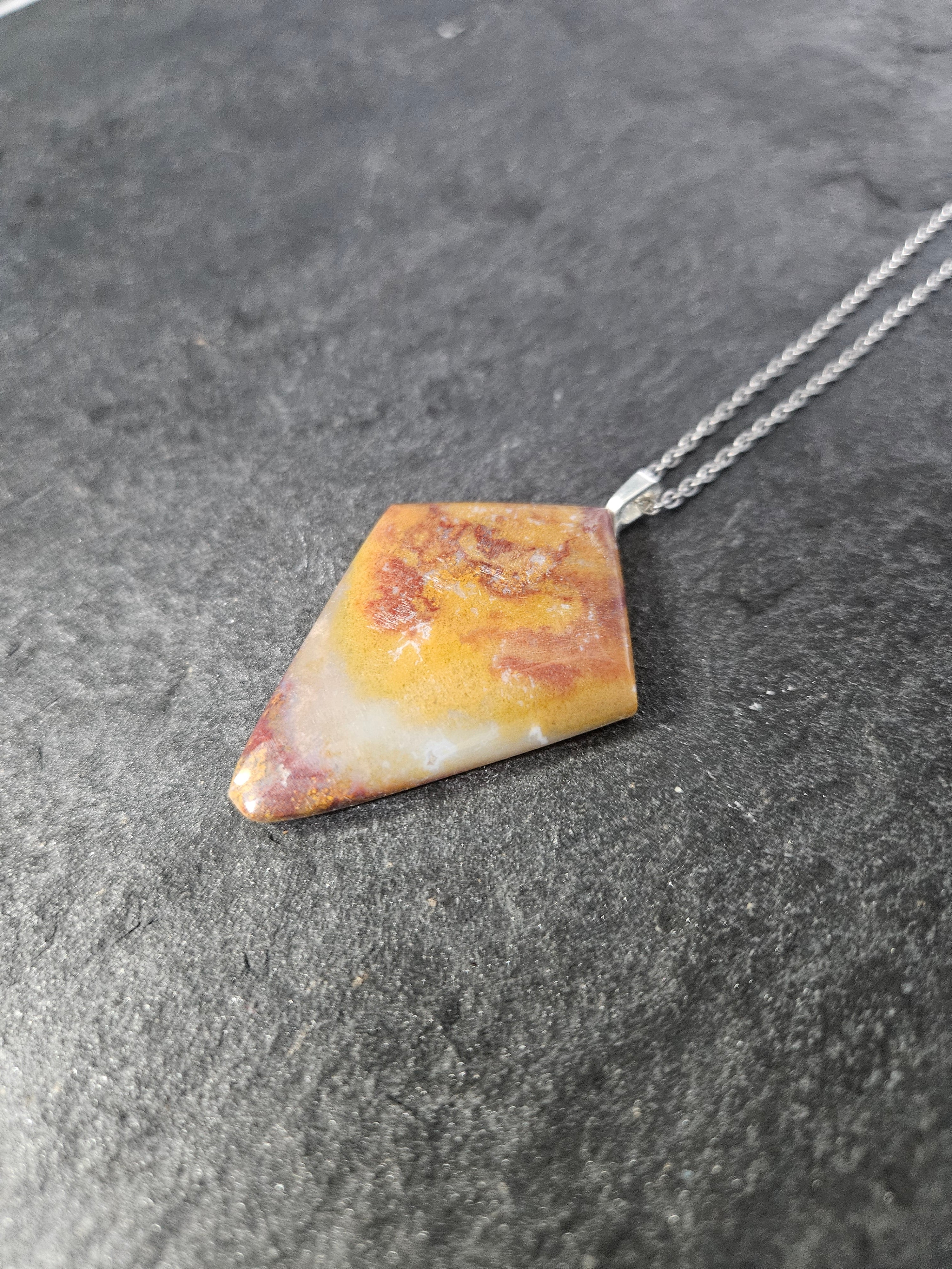 A geometric triangular pendant necklace made of marbled yellow, orange, and white stone with a silver chain, resting on a dark textured slate surface.