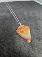 A triangular pendant necklace made from a marbled stone with yellow, orange, and white patterns, suspended on a silver chain over a dark gray textured surface.