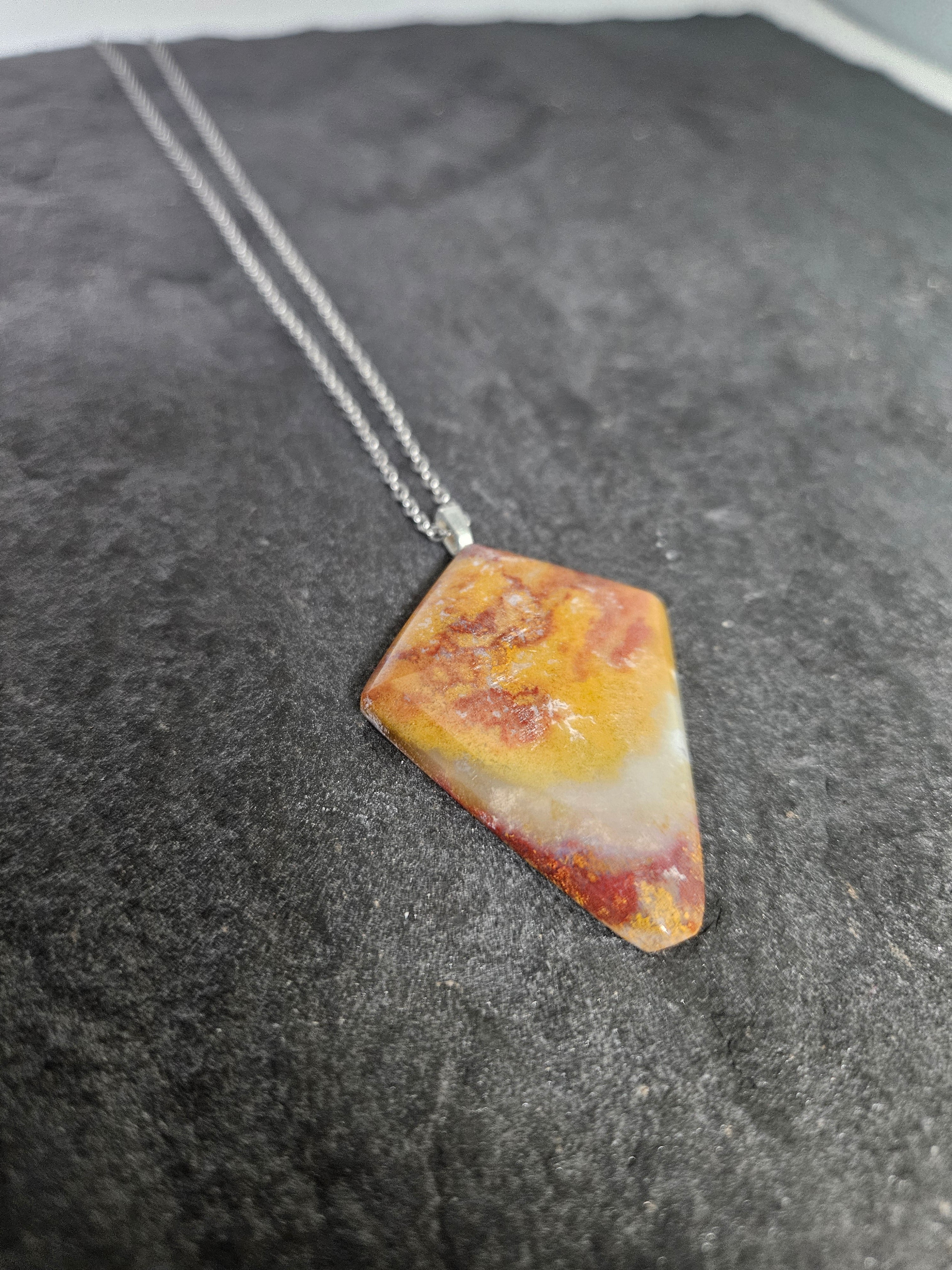 A triangular pendant necklace made from a marbled stone with yellow, orange, and white patterns, suspended on a silver chain over a dark gray textured surface.
