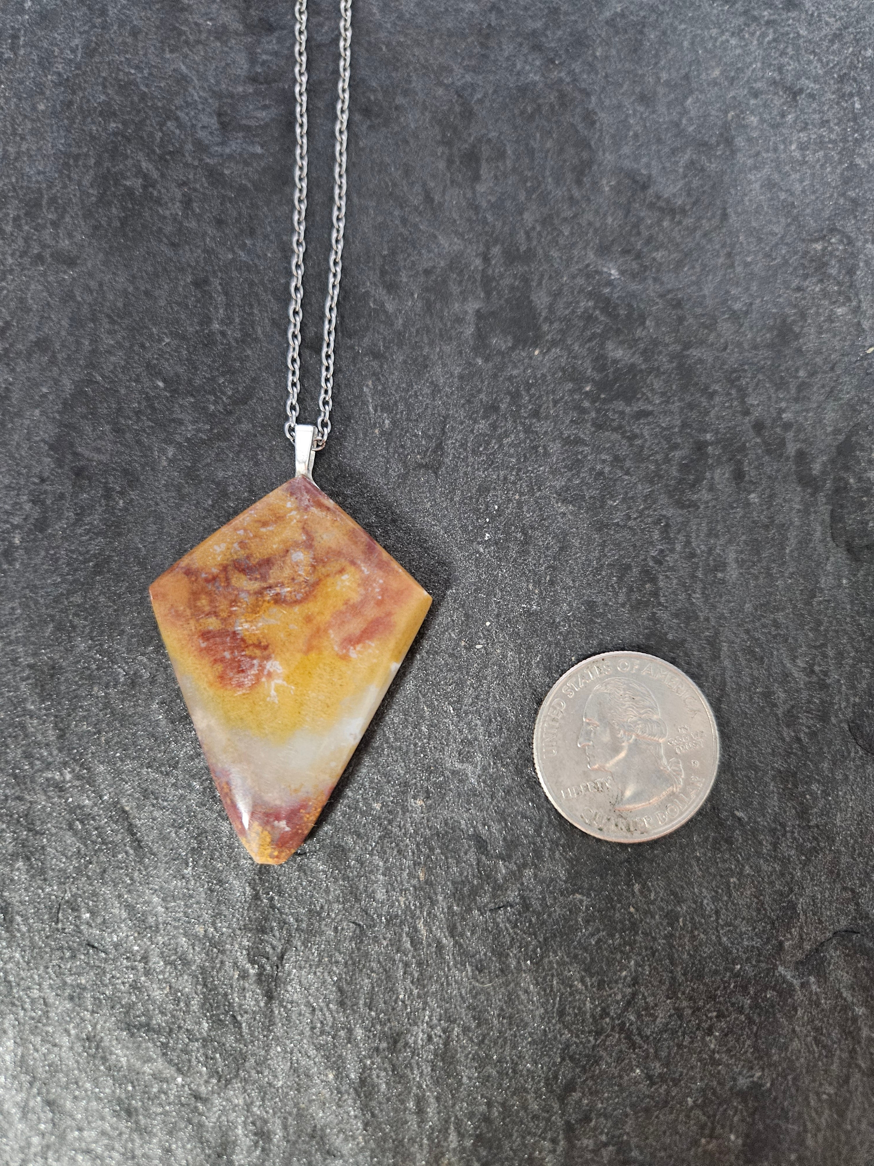 A diamond-shaped pendant necklace made of a marbled yellow, orange, and white stone on a silver chain, shown next to a US quarter coin for scale on a dark gray slate background.