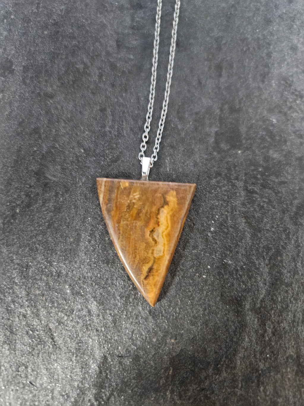 A triangular pendant necklace made of polished brown stone with natural banding, suspended from a silver chain, displayed on a dark textured surface.