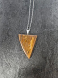 A triangular pendant necklace made of polished brown stone with natural banding, suspended from a silver chain, displayed on a dark textured surface.