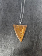A triangular pendant necklace made of polished brown stone with natural banding, suspended from a silver chain, displayed on a dark textured surface.