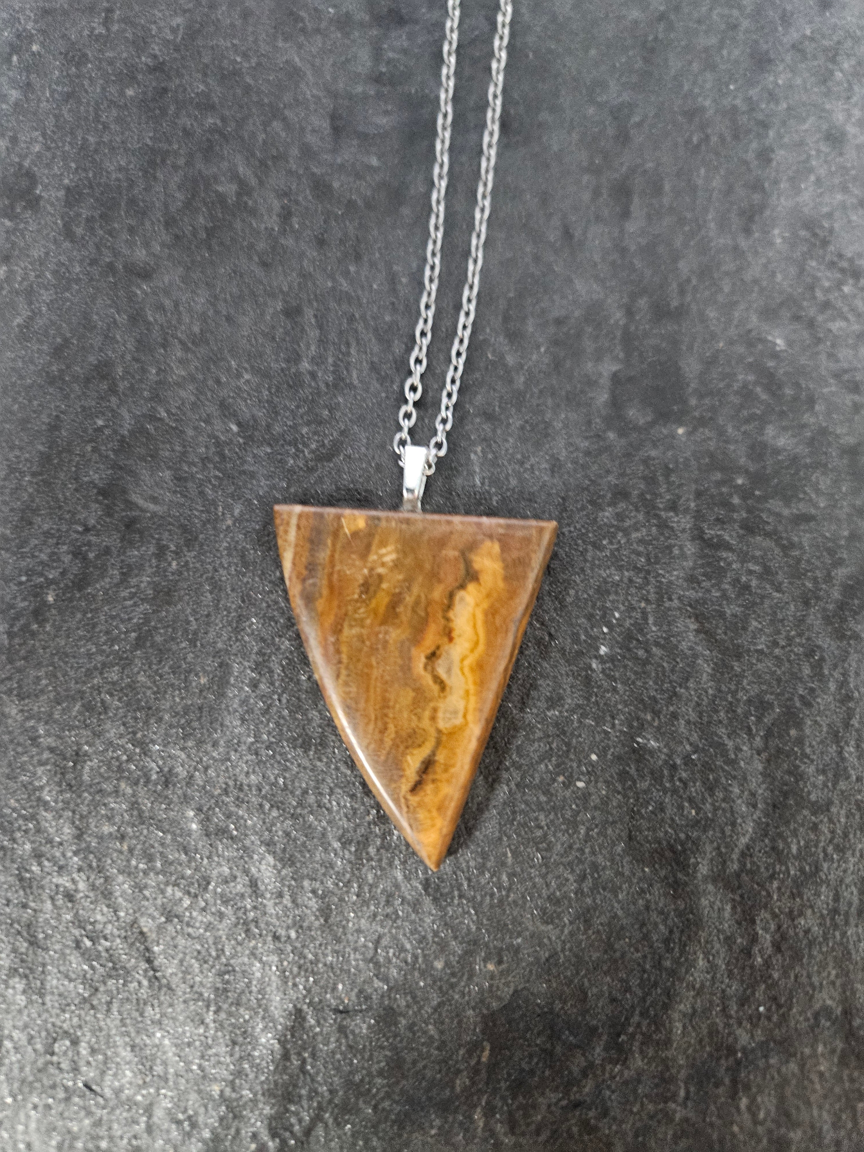 A triangular pendant necklace made of polished brown stone with natural banding, suspended from a silver chain, displayed on a dark textured surface.