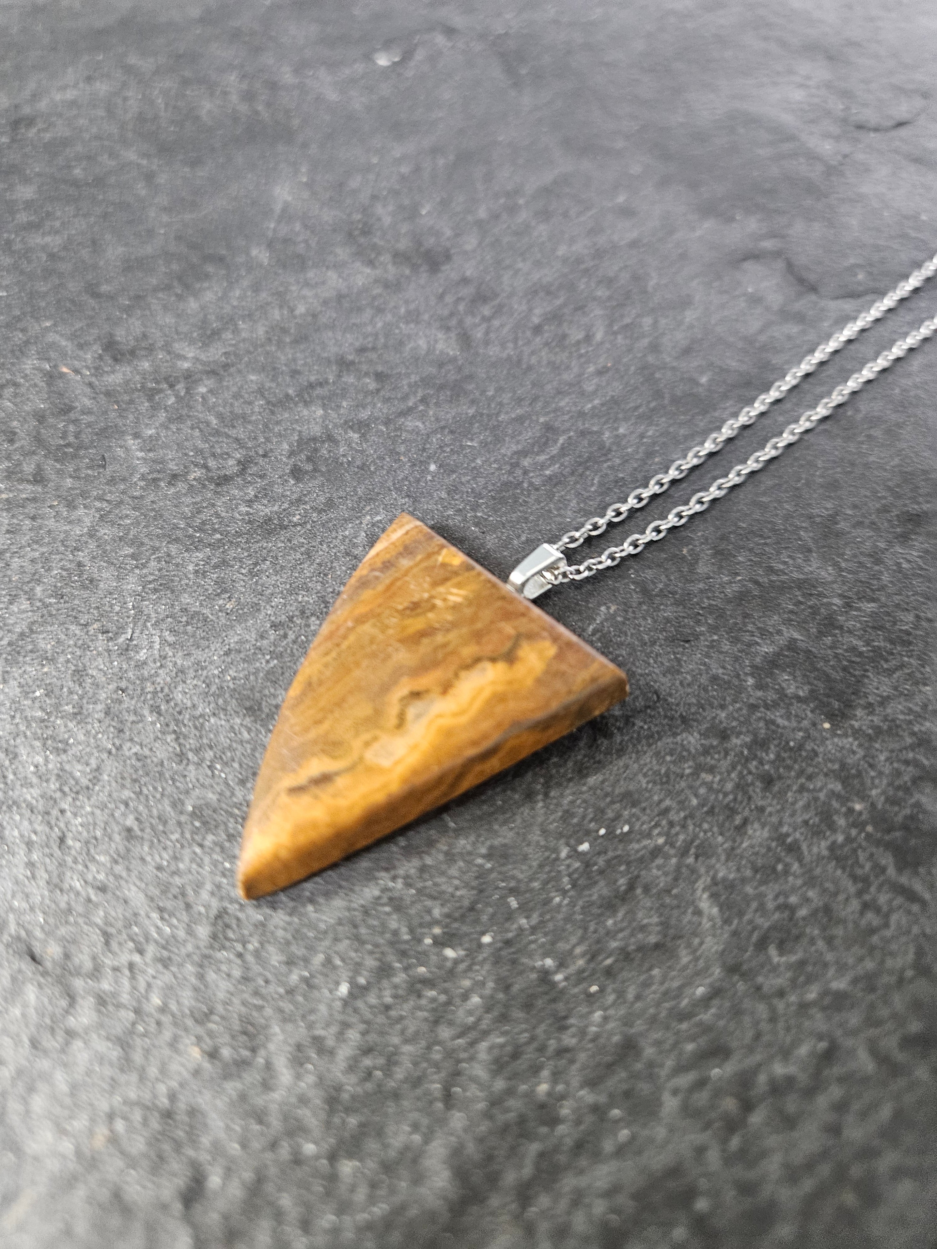 A triangular pendant necklace made from polished tiger's eye, hanging from a silver chain against a dark textured slate background.