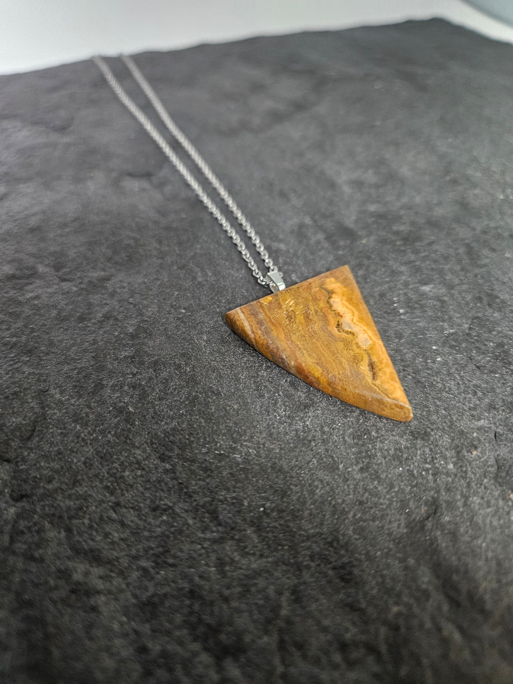 A triangular pendant made of natural brown wood or bone with visible grain, hanging from a silver chain against a dark textured background.
