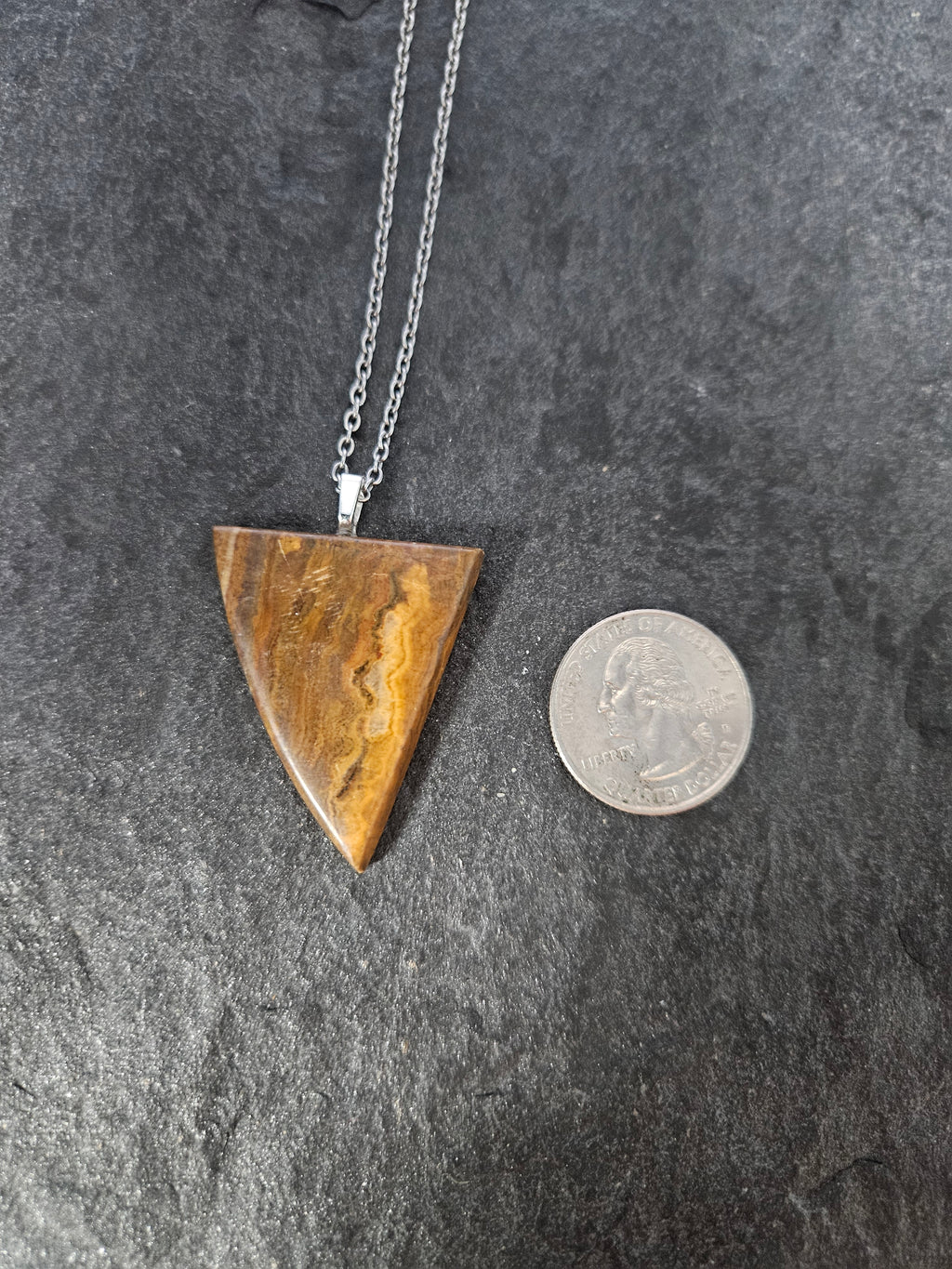 A triangular pendant necklace made of polished brown tiger's eye with natural banding, suspended from a silver chain, shown next to a US quarter coin for scale on a dark textured slate background.