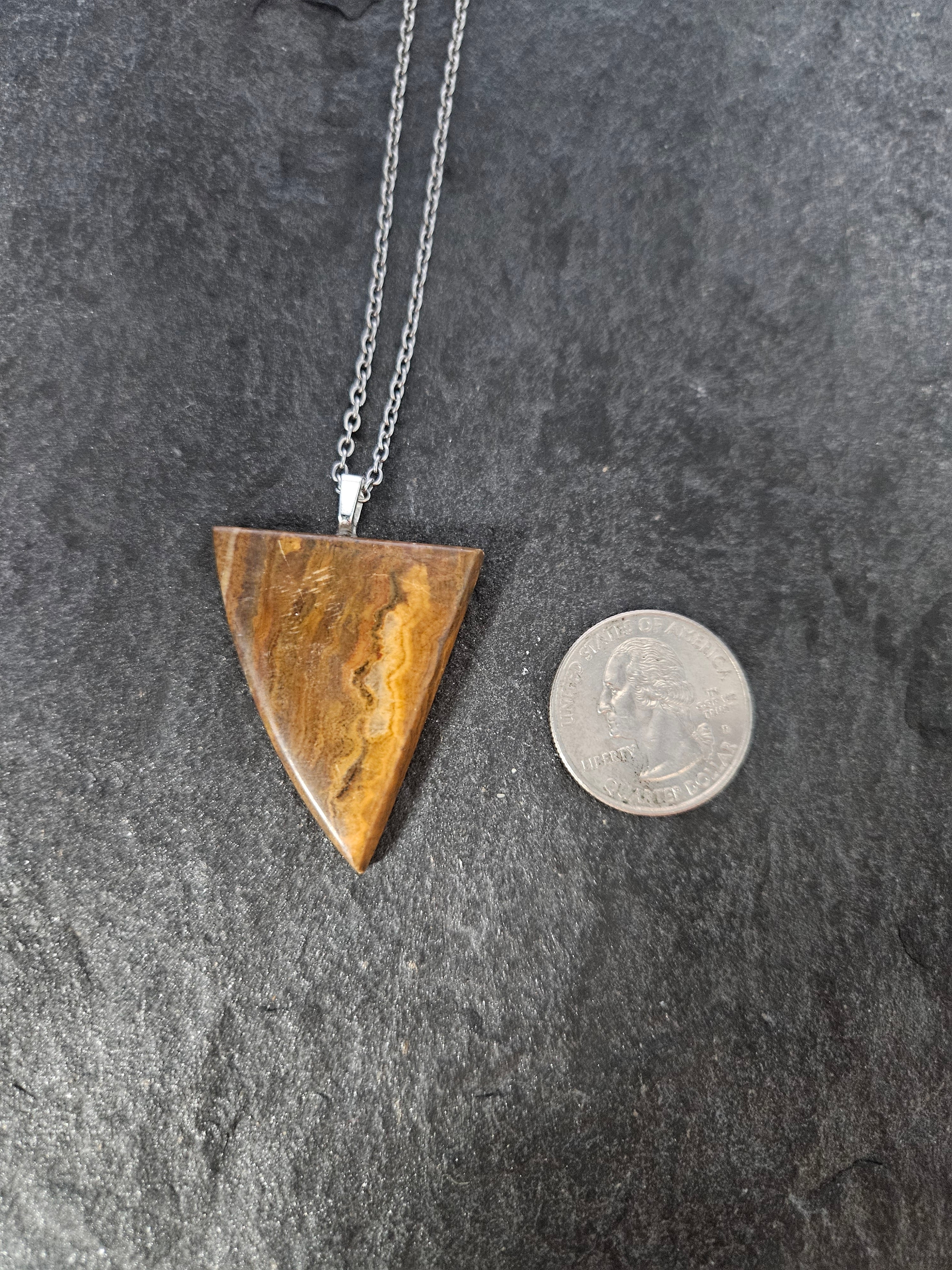 A triangular pendant necklace made of polished brown tiger's eye with natural banding, suspended from a silver chain, shown next to a US quarter coin for scale on a dark textured slate background.