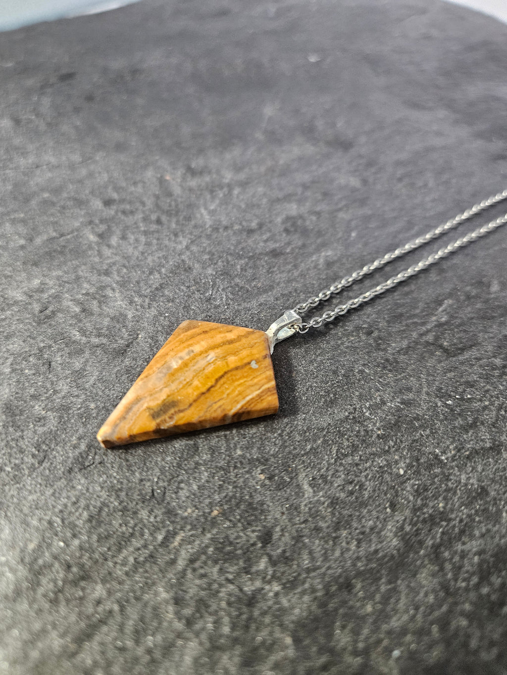 A geometric pendant necklace featuring a polished triangular piece of tiger's eye stone with natural brown and orange banding, suspended from a silver-toned chain and resting on a dark textured surface.