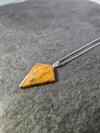 A geometric pendant necklace featuring a polished triangular piece of tiger's eye stone with natural brown and orange banding, suspended from a silver-toned chain and resting on a dark textured surface.