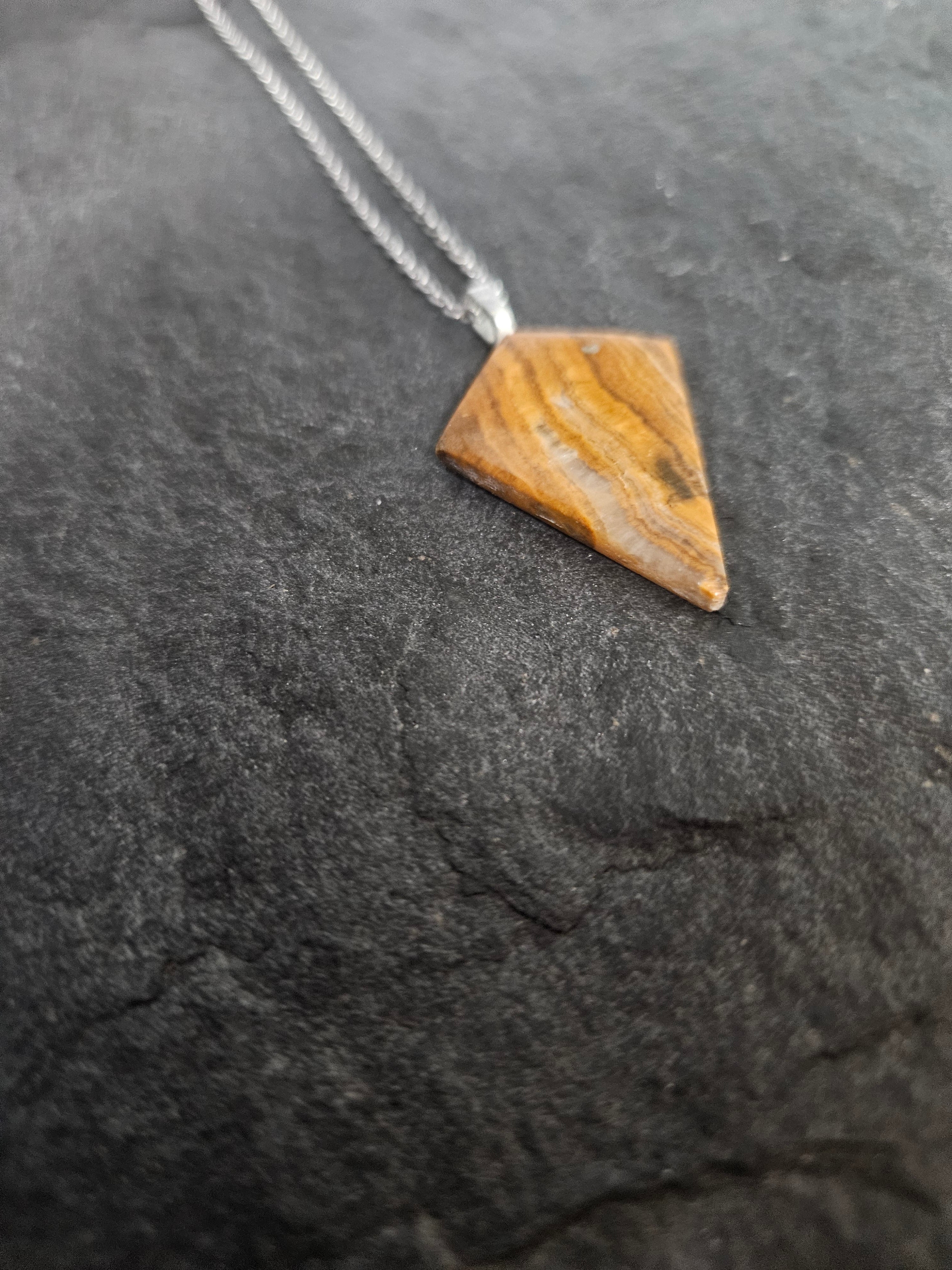 A triangular wooden pendant with natural brown and tan grain, suspended from a silver-colored chain, displayed on a dark textured stone surface.
