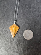 A triangular pendant necklace made of polished brown and gold striped stone, hanging from a silver chain on a dark textured surface, with a coin placed above it for scale.