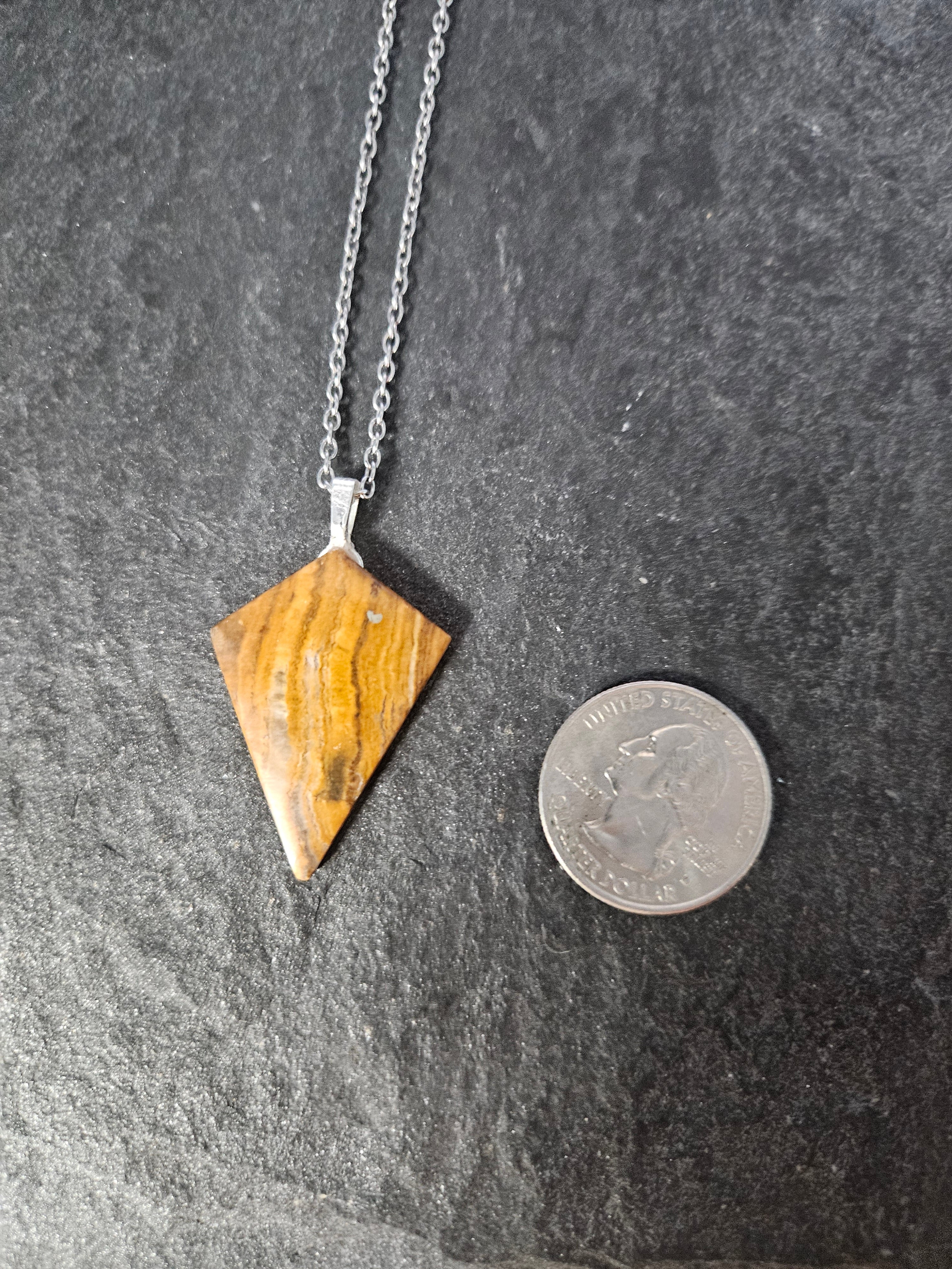 A triangular pendant necklace made of polished brown and gold striped stone, hanging from a silver chain on a dark textured surface, with a coin placed above it for scale.