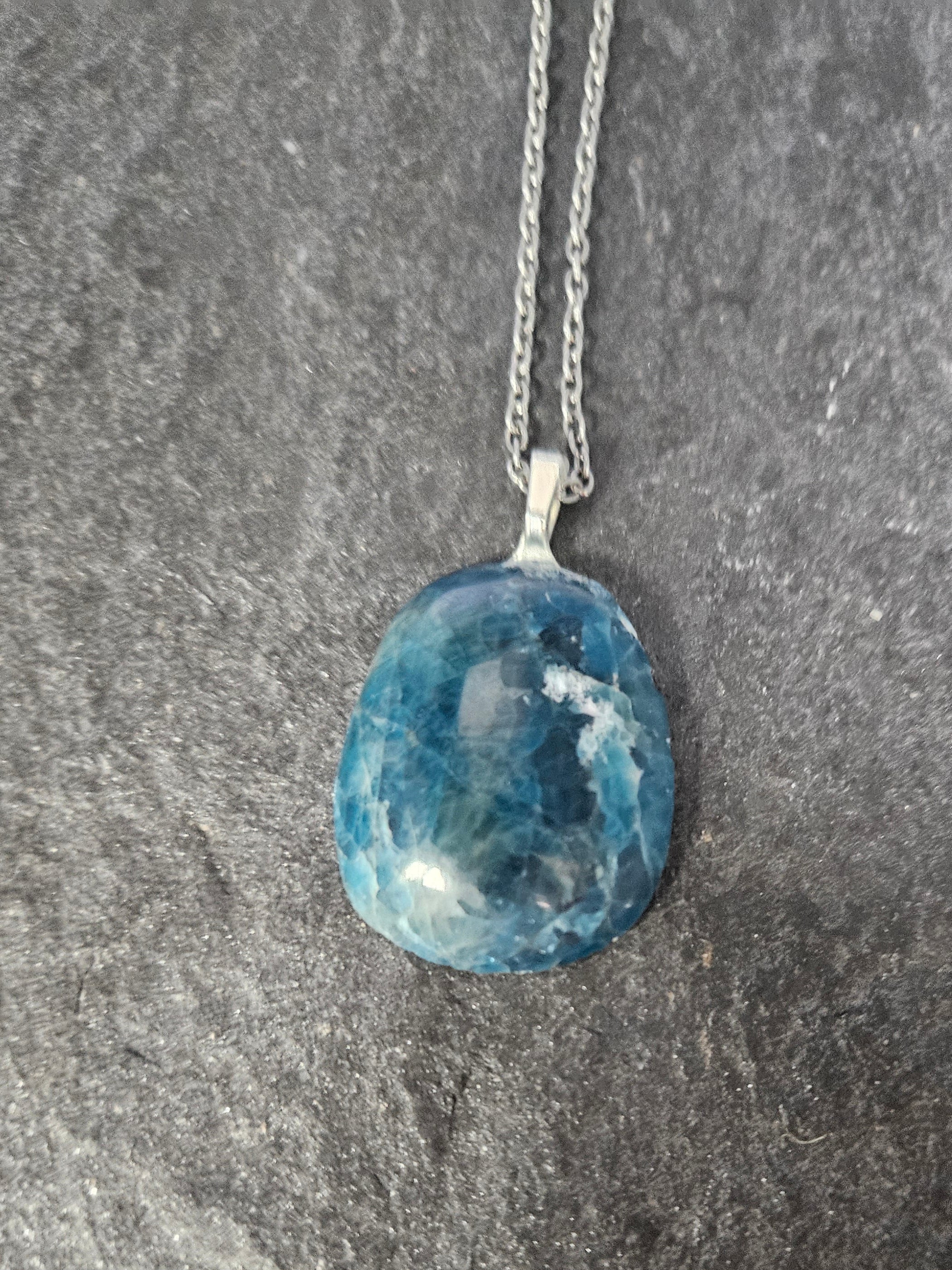 A blue marbled gemstone pendant on a silver chain, displayed against a dark textured slate background.