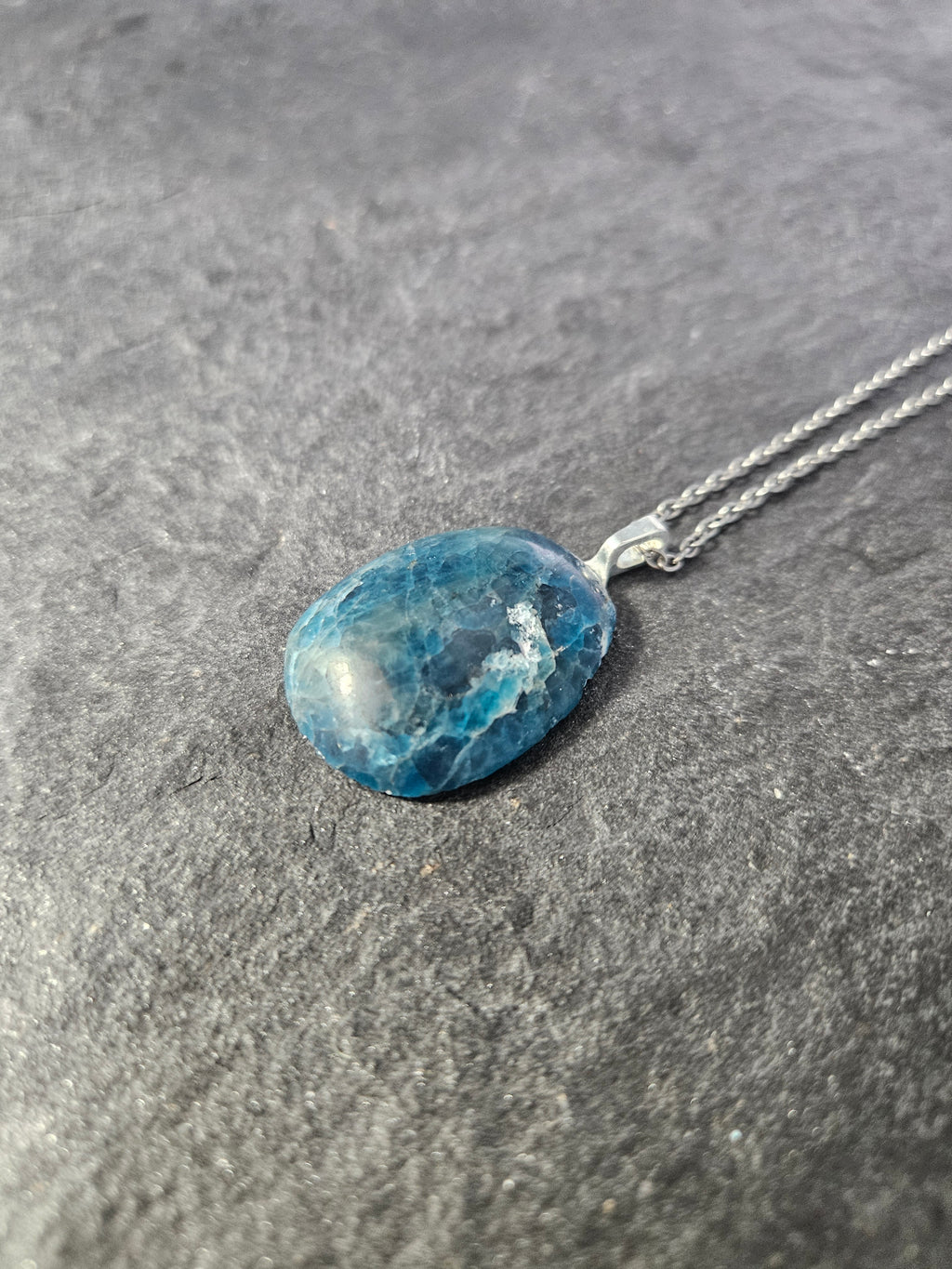 A silver chain necklace with an oval blue marbled stone pendant, displayed on a textured gray surface.