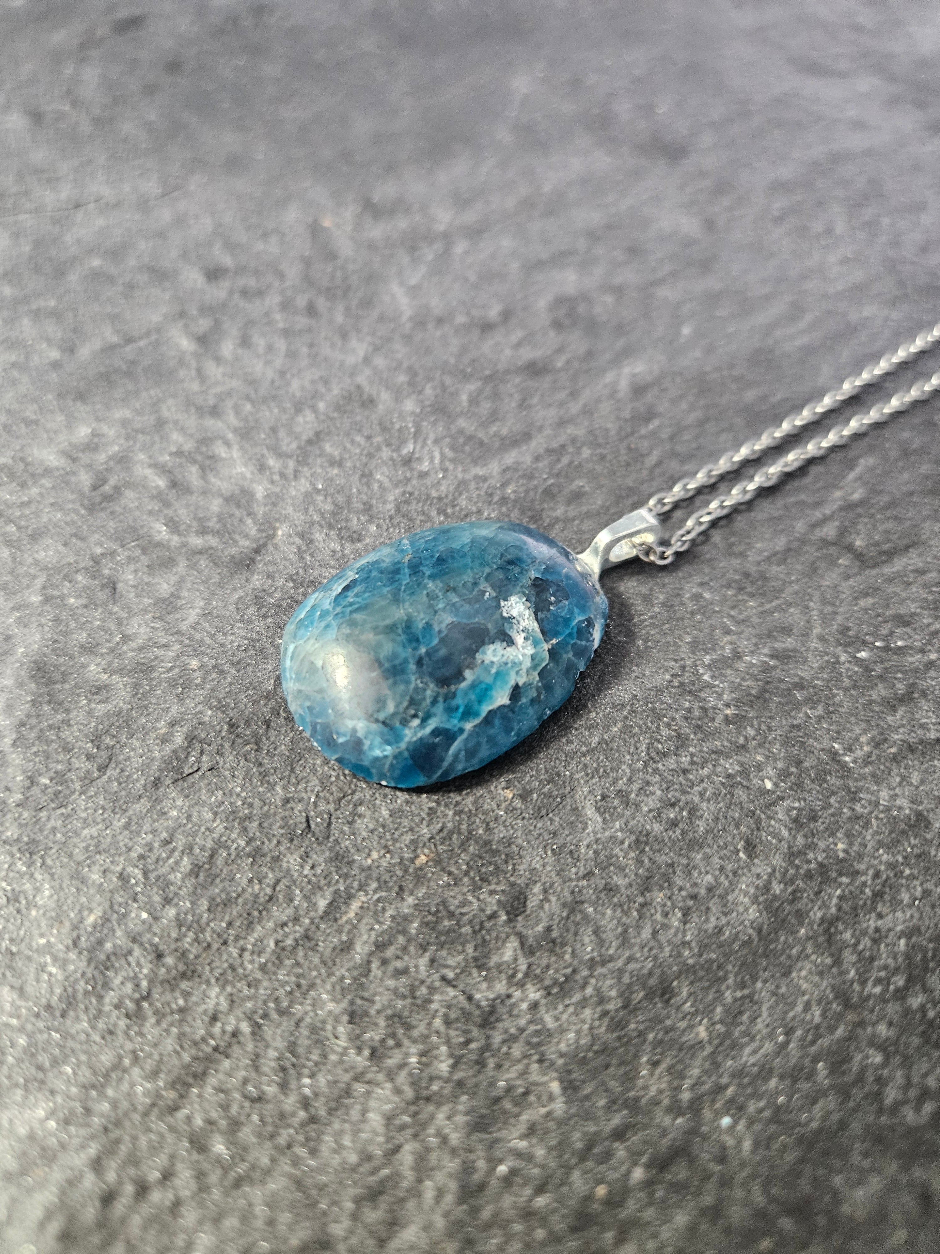 A silver chain necklace with an oval blue marbled stone pendant, displayed on a textured gray surface.