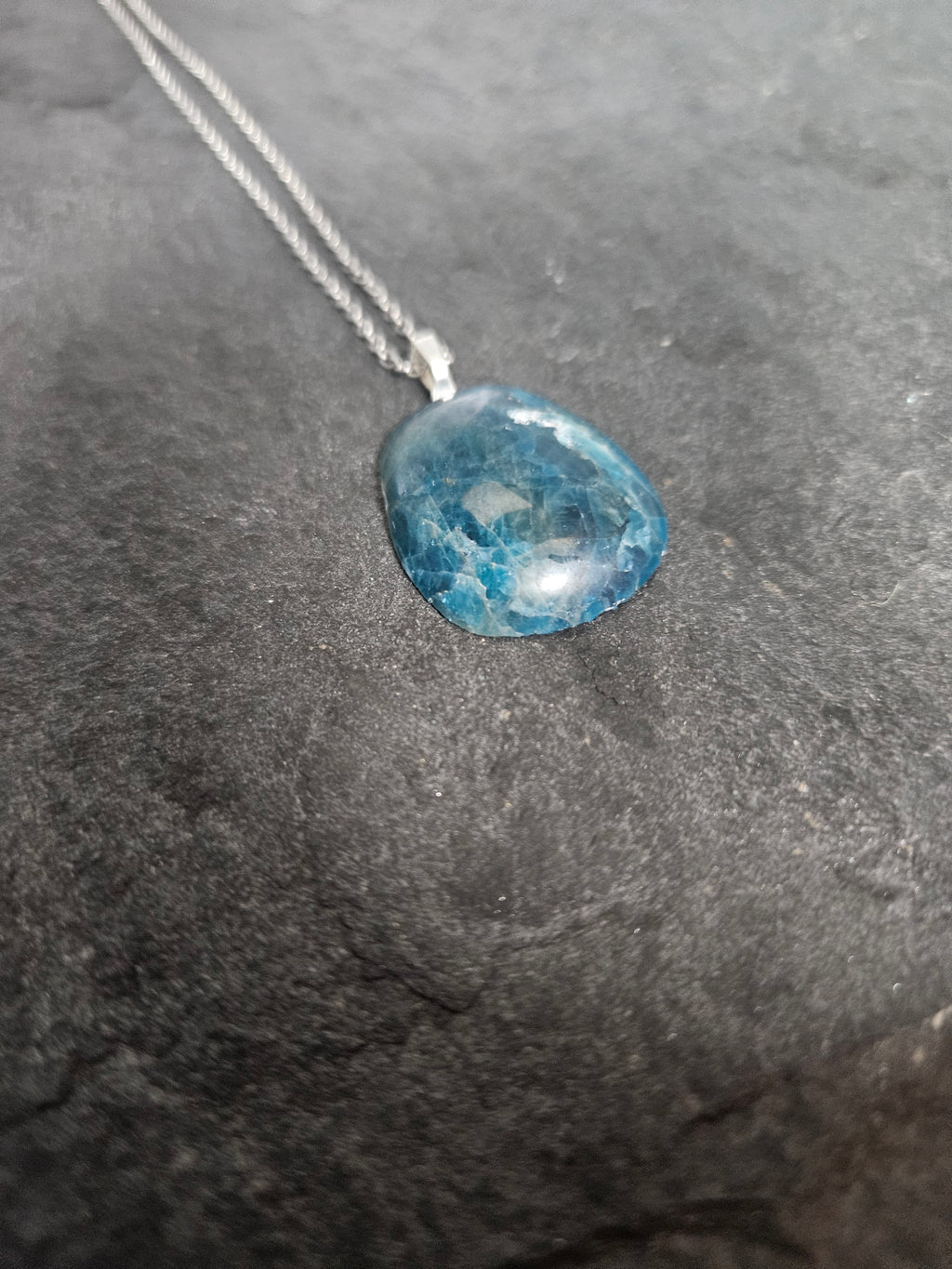 A silver necklace with a polished, irregular-shaped blue gemstone pendant, resting on a dark textured slate surface.