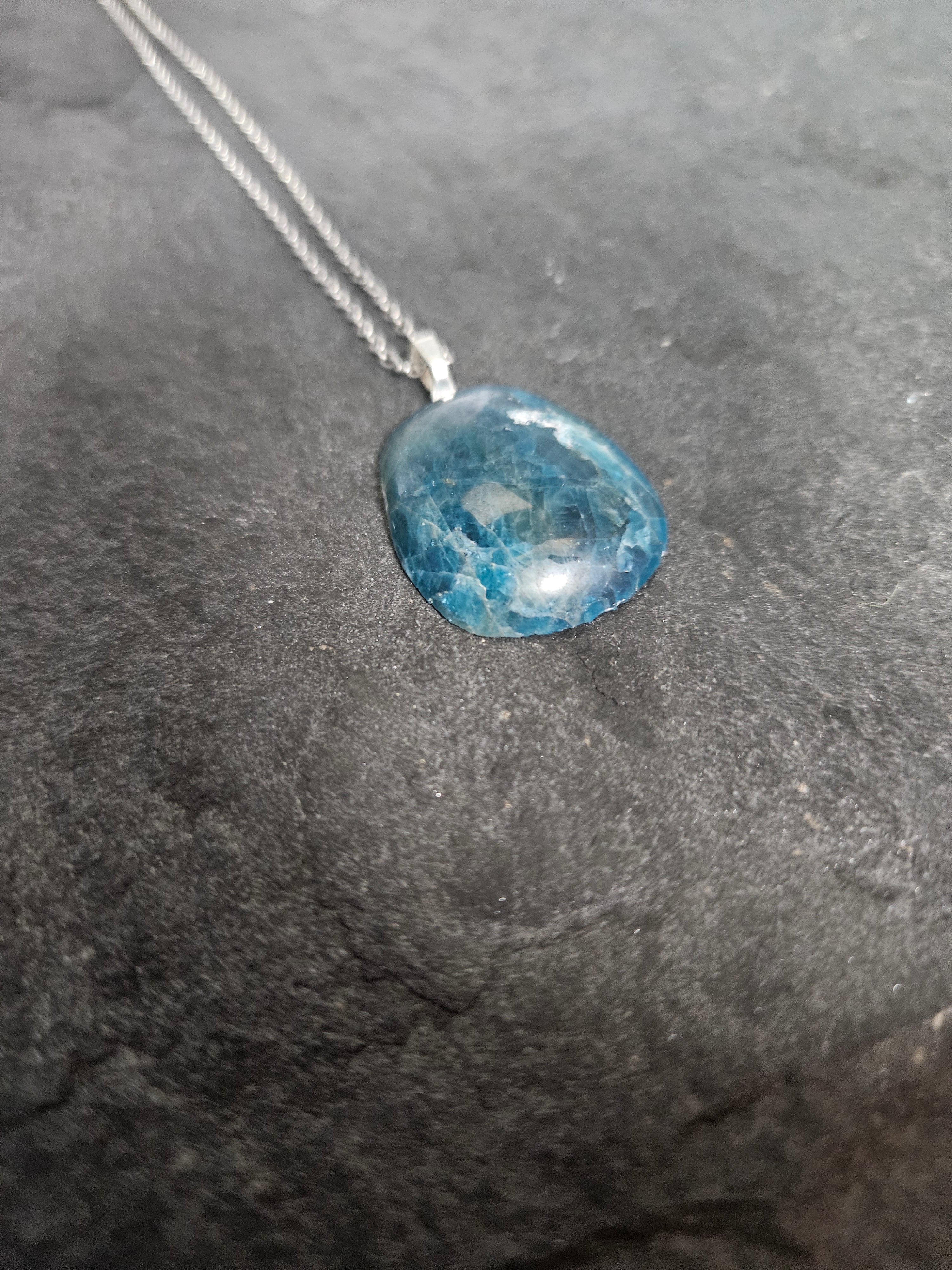 A silver necklace with a polished, irregular-shaped blue gemstone pendant, resting on a dark textured slate surface.