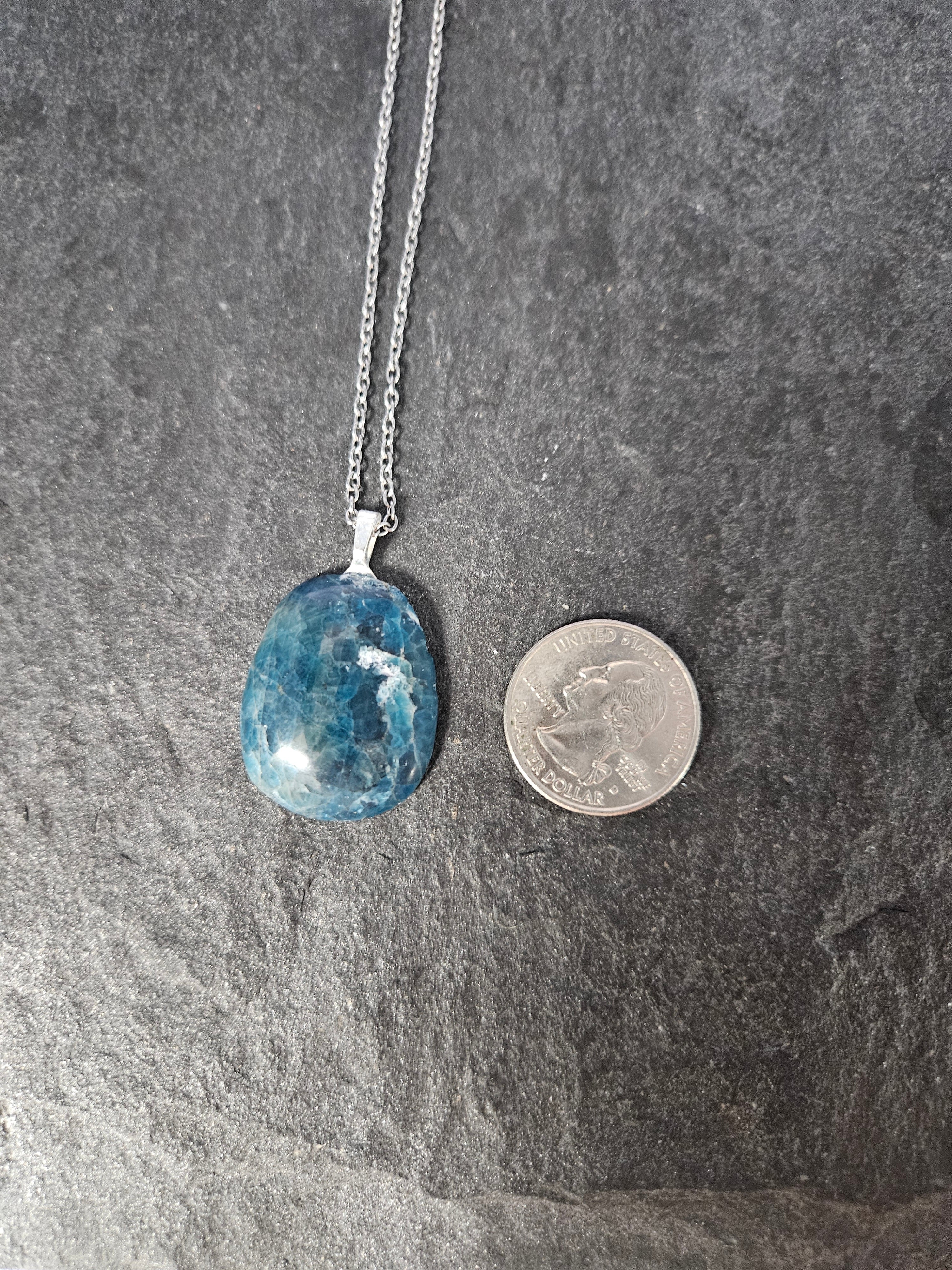 A blue teardrop-shaped gemstone pendant necklace on a silver chain, displayed next to a US quarter coin on a dark gray slate surface.