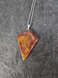 A geometric pendant necklace featuring a triangular stone with a marbled pattern of orange, yellow, and reddish-brown, suspended from a silver chain against a dark gray slate background.