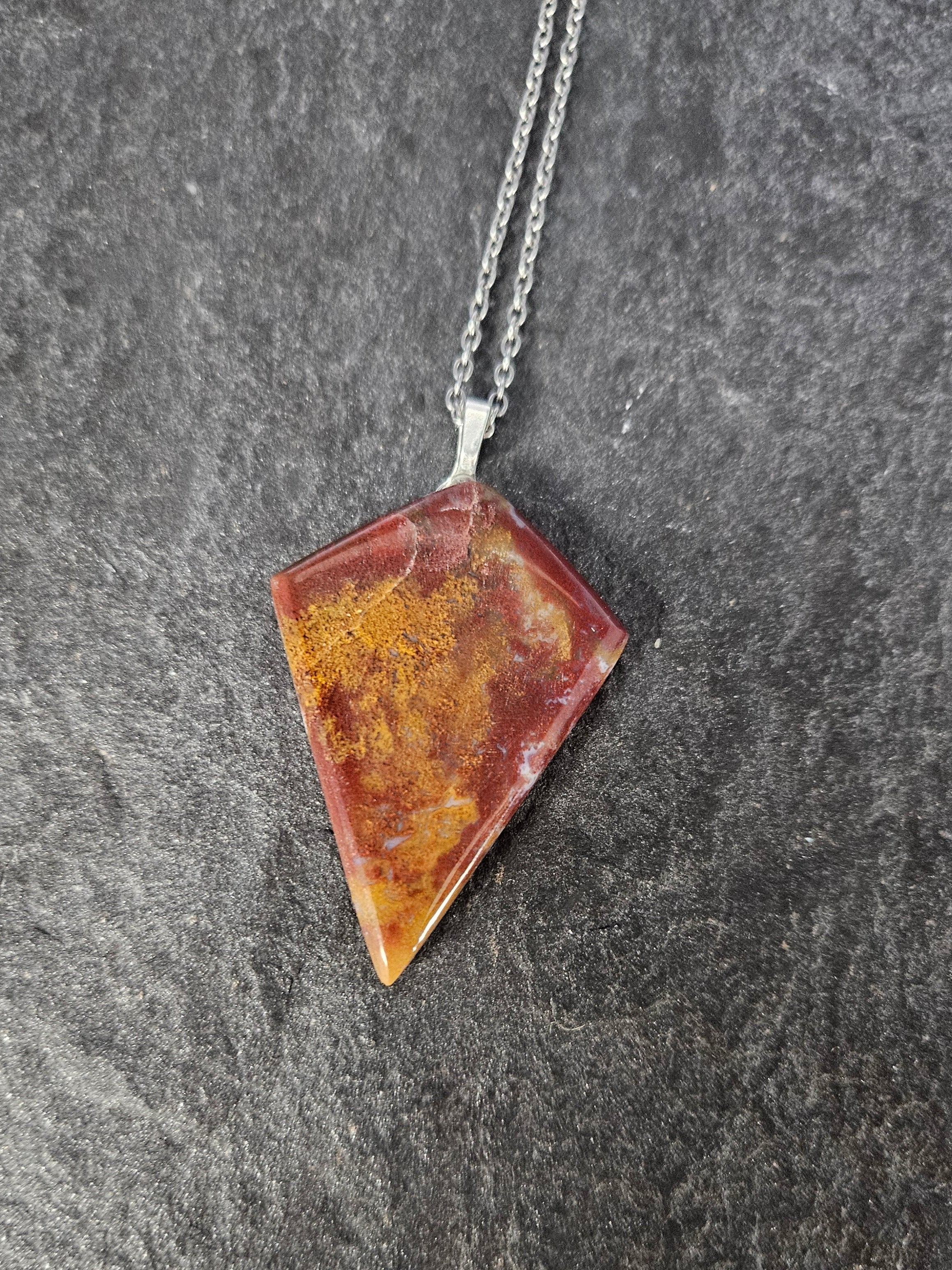 A geometric pendant necklace featuring a triangular stone with a marbled pattern of orange, yellow, and reddish-brown, suspended from a silver chain against a dark gray slate background.