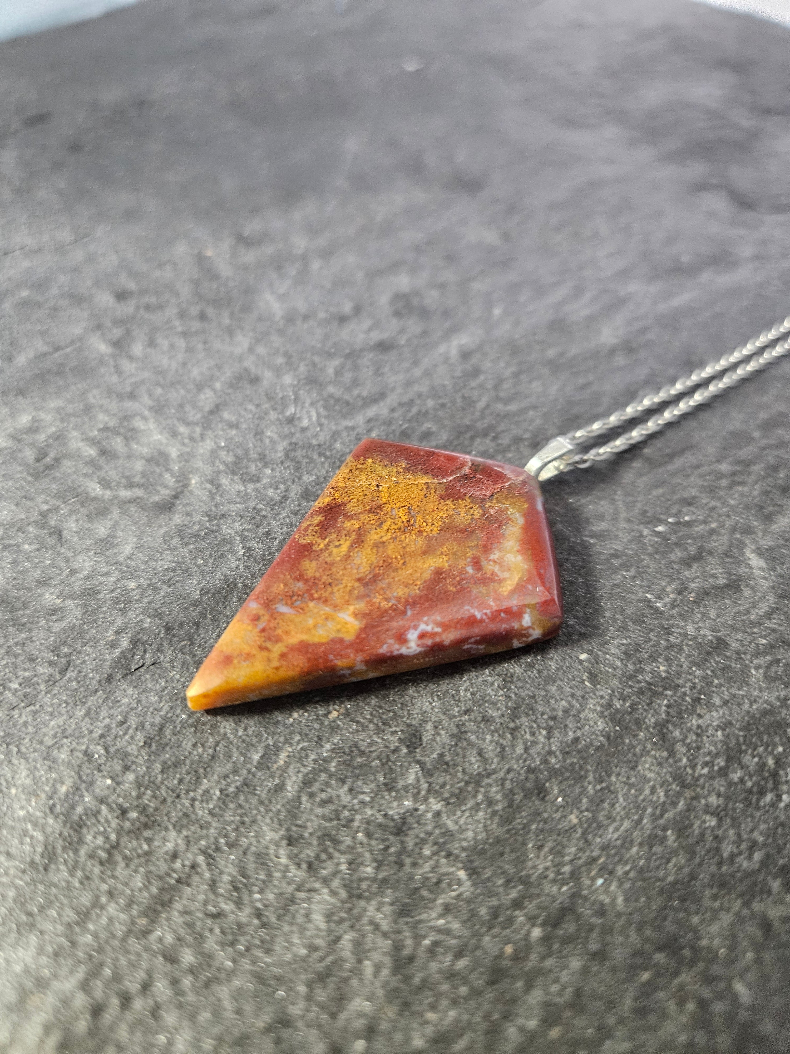 A triangular pendant necklace made of a marbled red and orange stone with gold flecks, attached to a silver chain, resting on a dark textured surface.