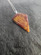 A triangular pendant necklace made of polished red and orange stone with a silver chain, resting on a dark textured slate surface.