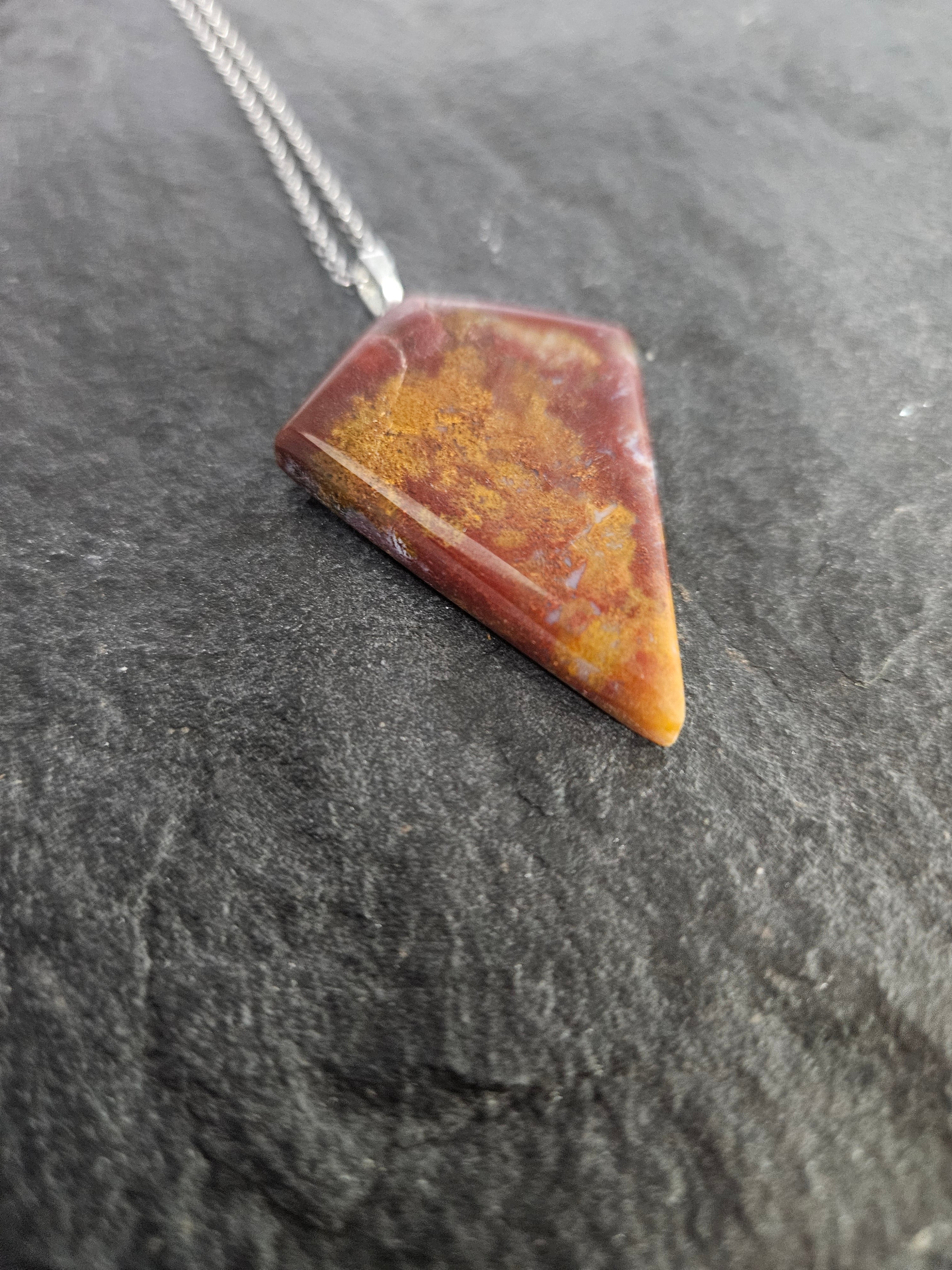 A triangular pendant necklace made of polished red and orange stone with a silver chain, resting on a dark textured slate surface.