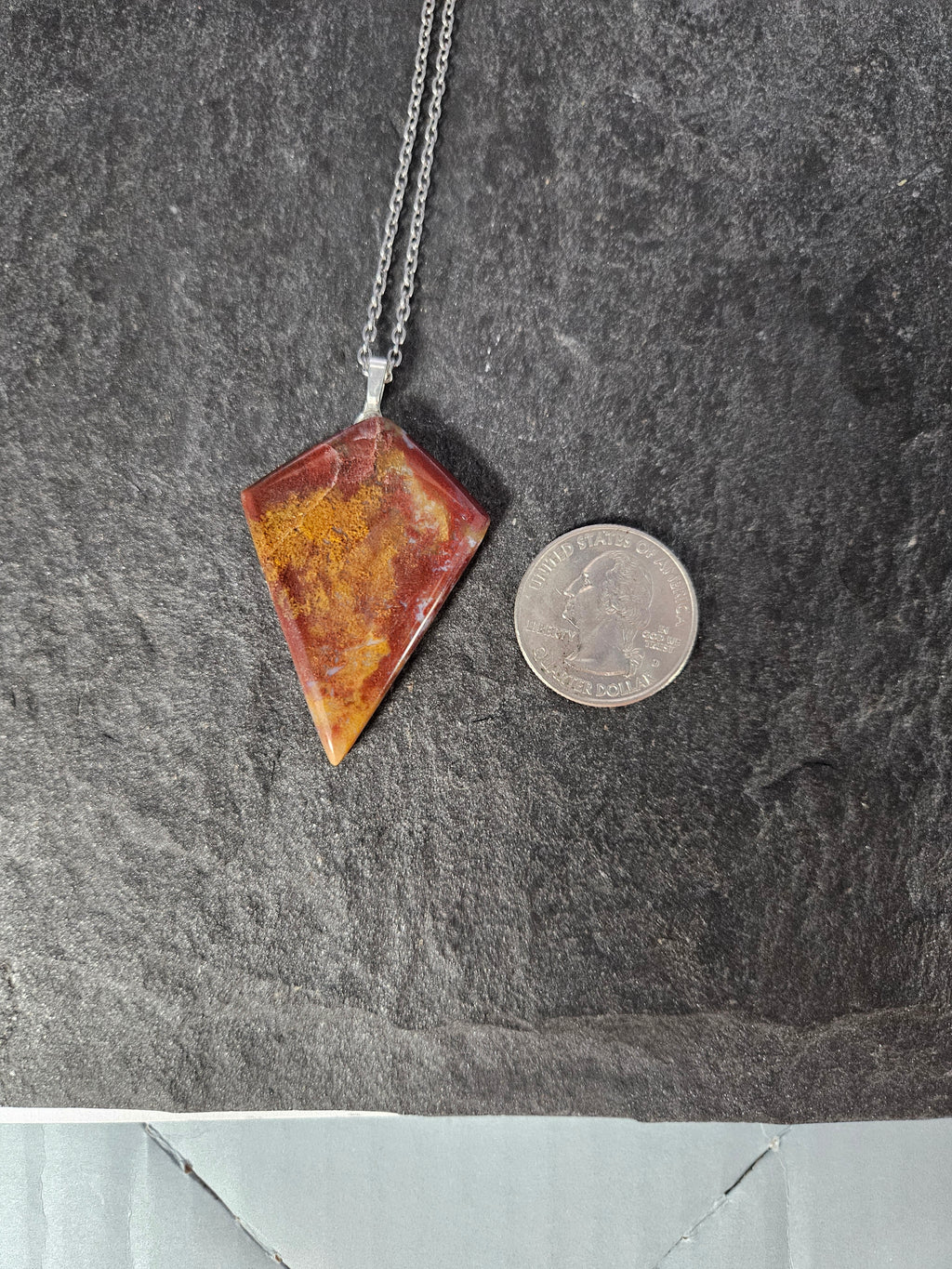 A silver chain necklace with a triangular pendant made of marbled red, orange, and yellow stone, shown next to a US quarter coin for scale on a dark gray slate surface.