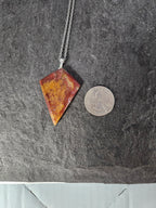 A silver chain necklace with a triangular pendant made of marbled red, orange, and yellow stone, shown next to a US quarter coin for scale on a dark gray slate surface.