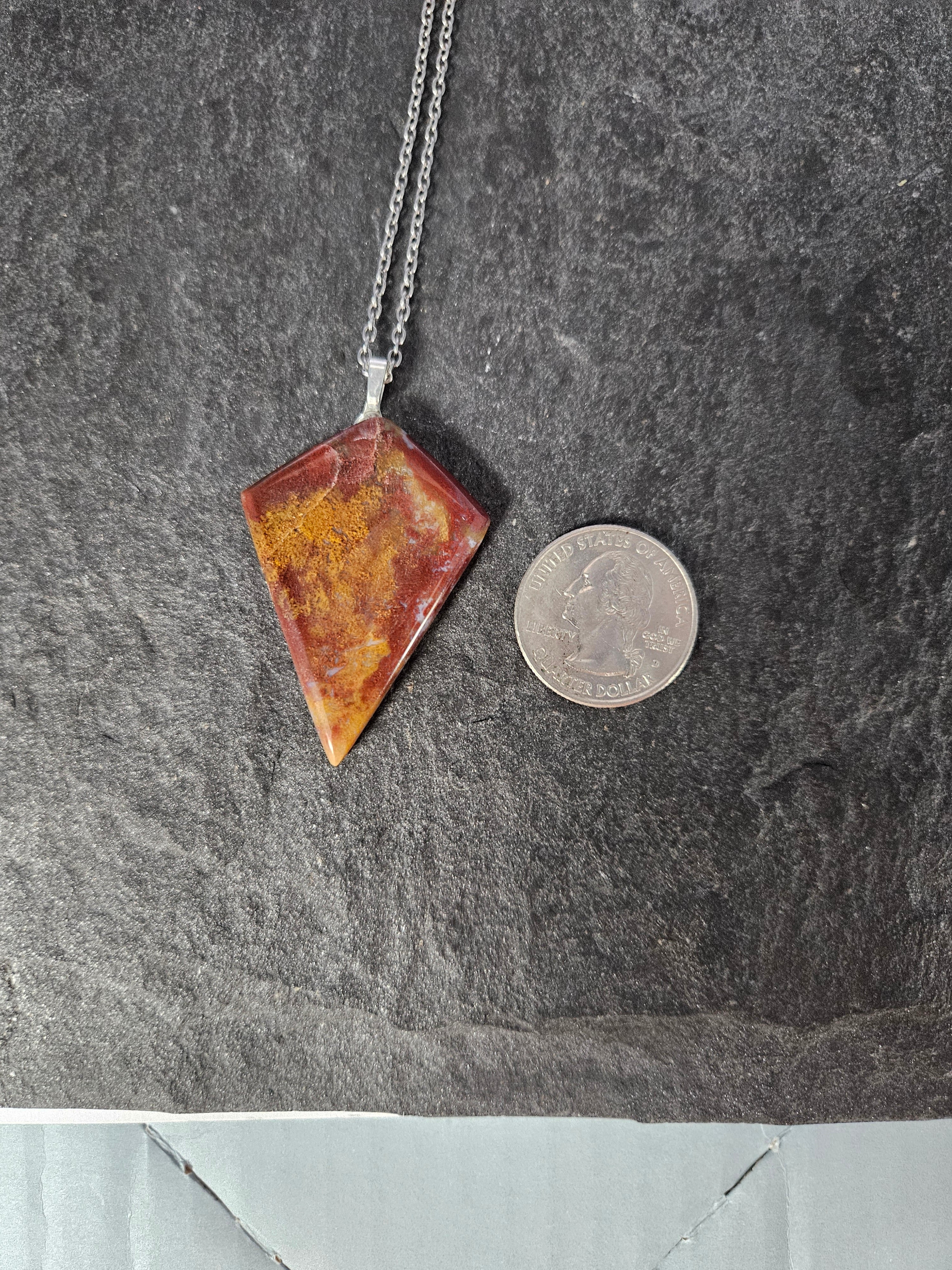 A silver chain necklace with a triangular pendant made of marbled red, orange, and yellow stone, shown next to a US quarter coin for scale on a dark gray slate surface.